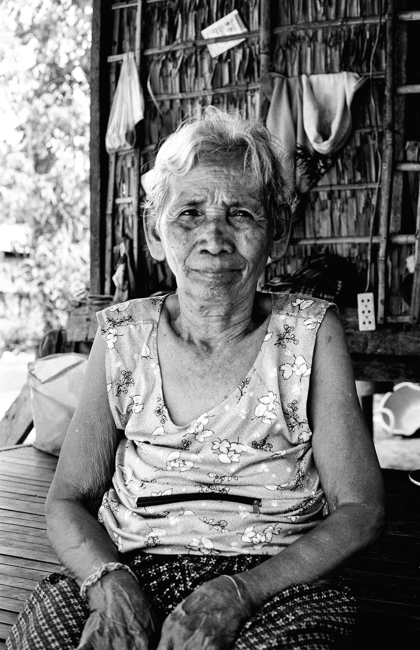 Elderly woman in floral dress sitting on wooden porch with baskets behind