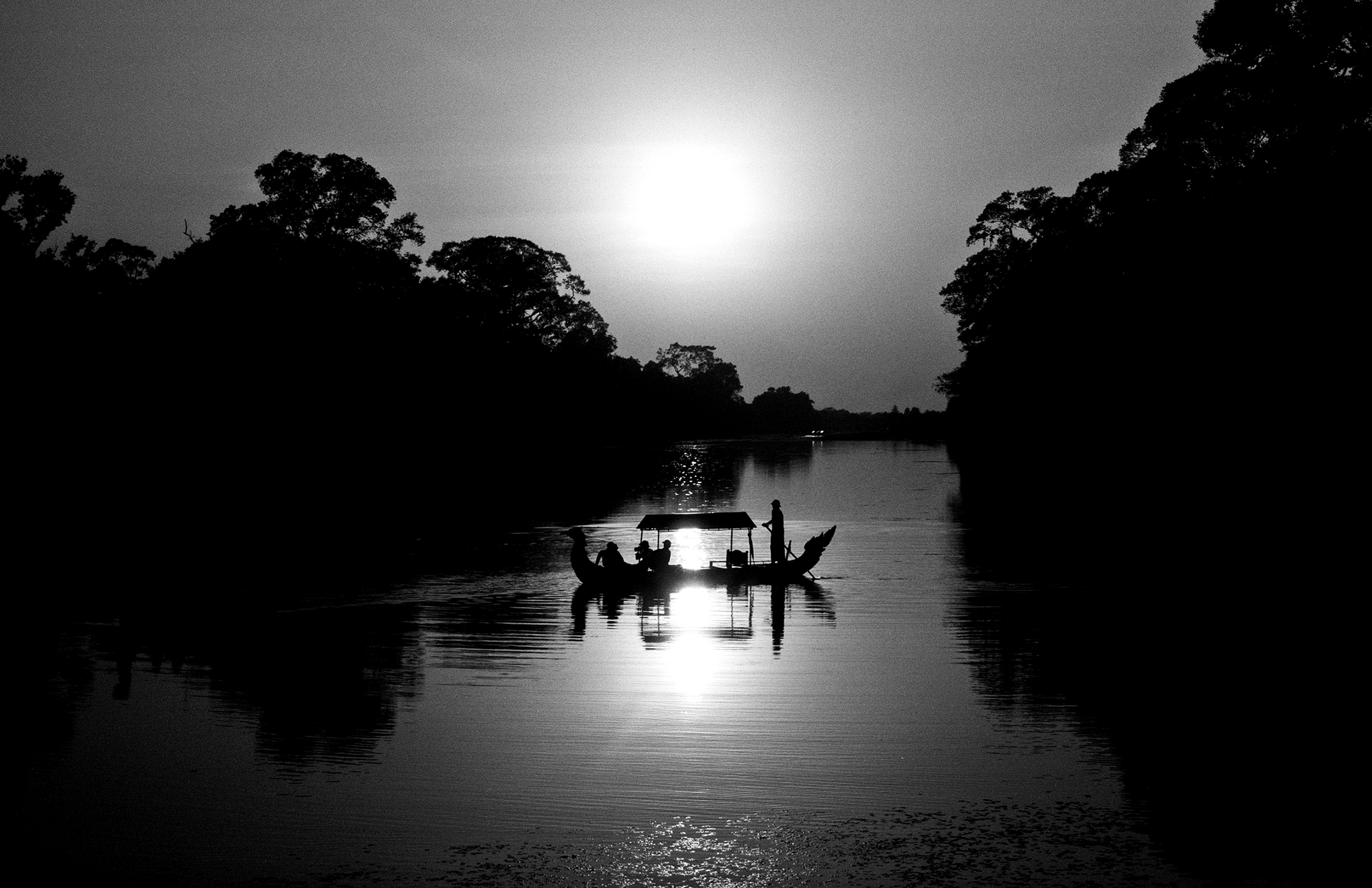 Silhouetted boat with passengers crossing illuminated river at dusk with forest backdrop
