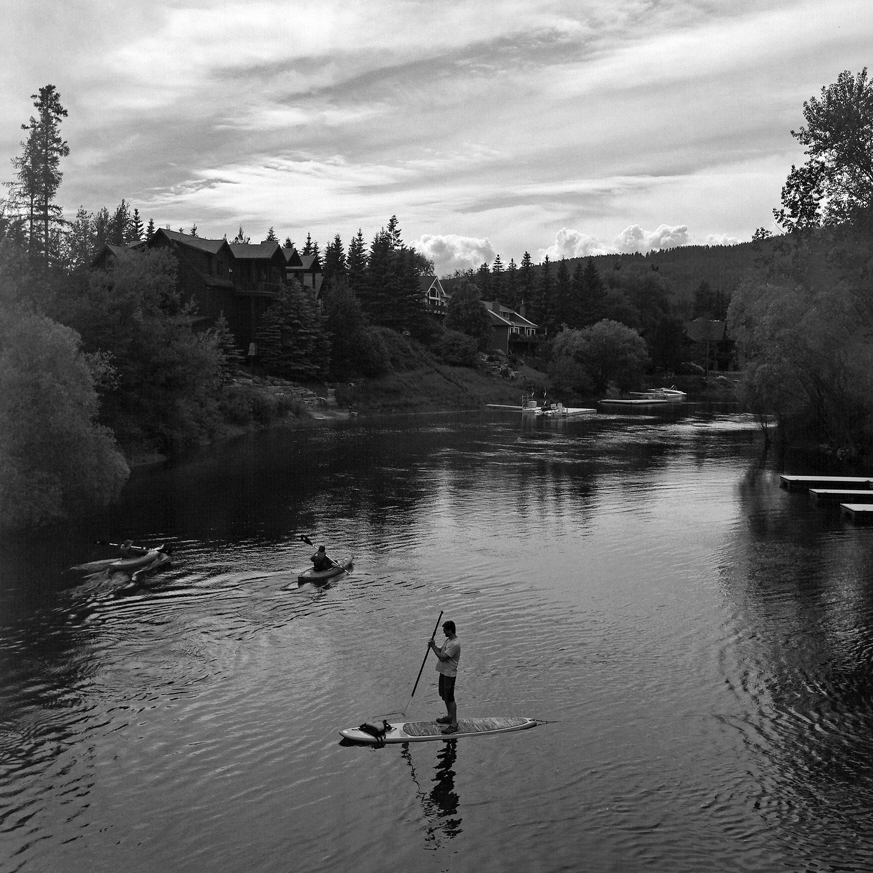 Paddleboarder and kayakers on calm lake with lakefront homes and forested hills under cloudy sky