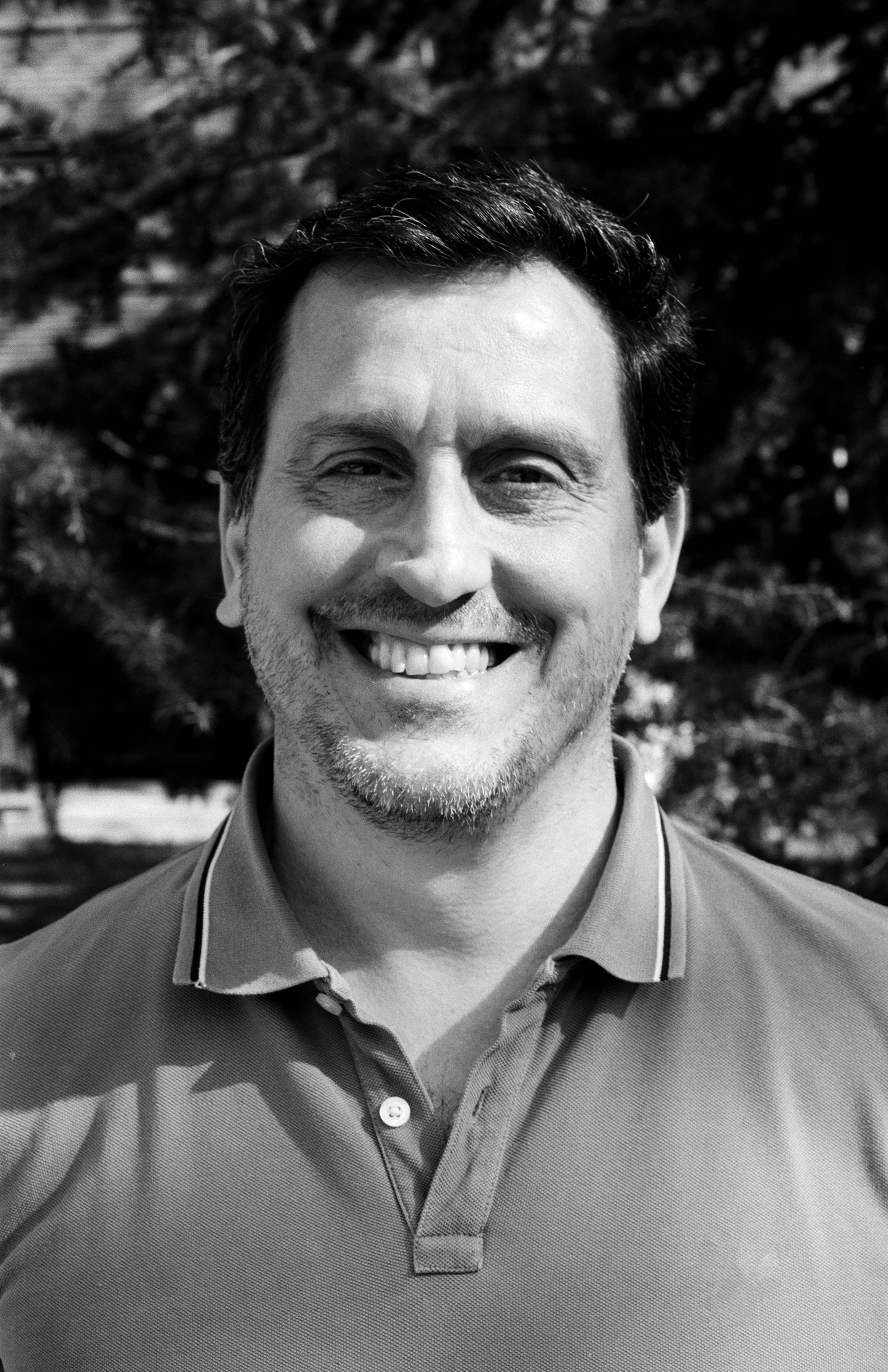 Portrait of middle-aged man smiling at camera wearing polo shirt against foliage background