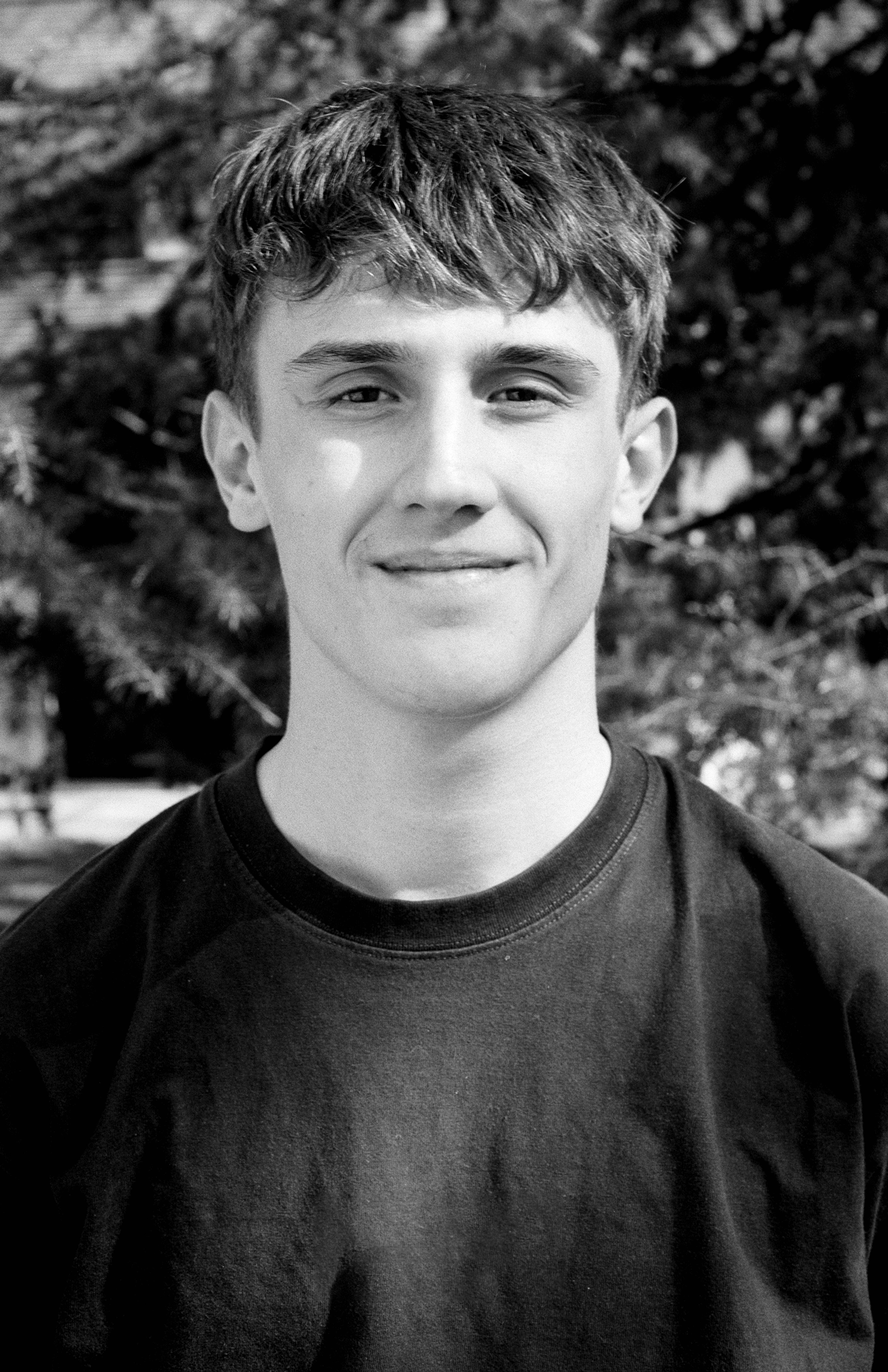 Portrait of young man smiling at camera against blurred foliage background in outdoor setting