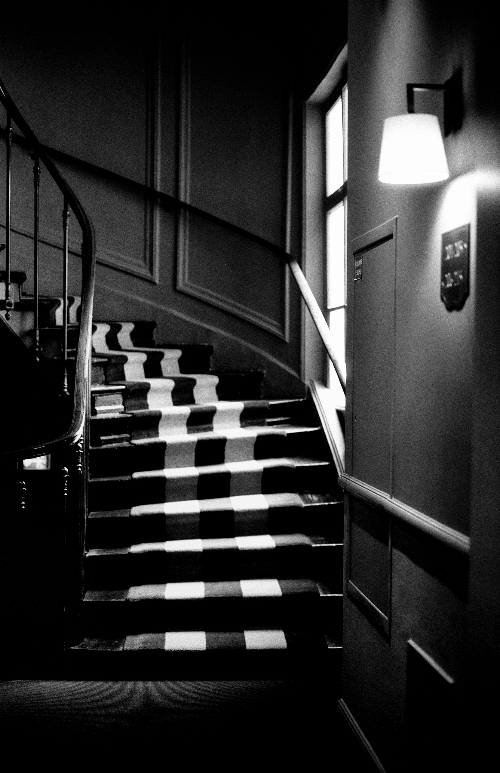 Ornate checkered staircase with curved wooden banister lit by wall lamp in dimly lit interior