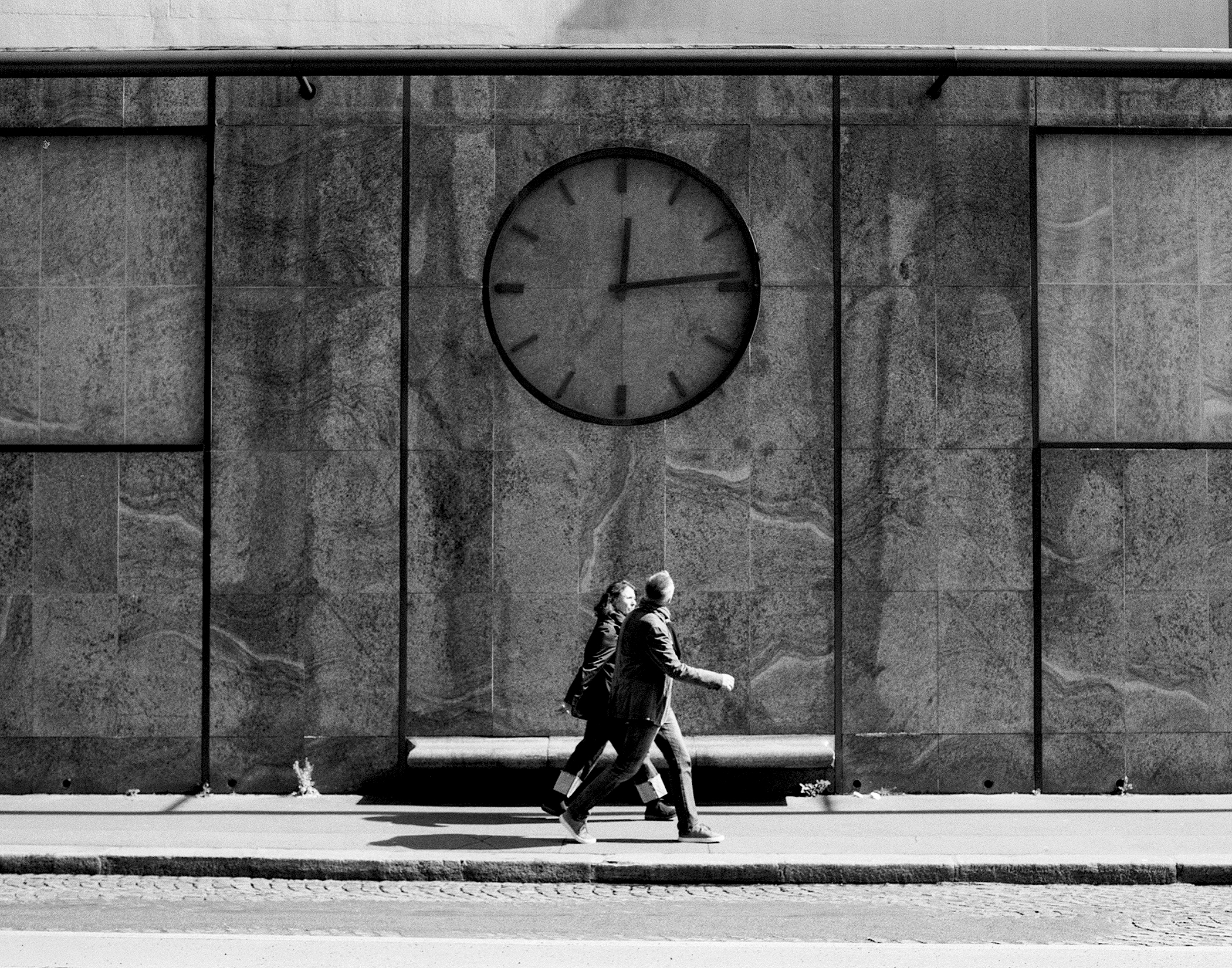 Two figures walking past large modernist clock face on grey stone wall
