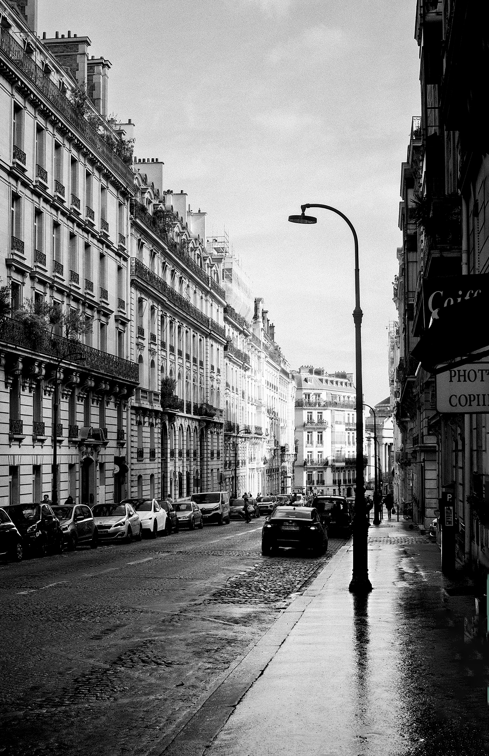 Wet Parisian street lined with classical Haussmann buildings, street lamp and parked cars reflected in rain