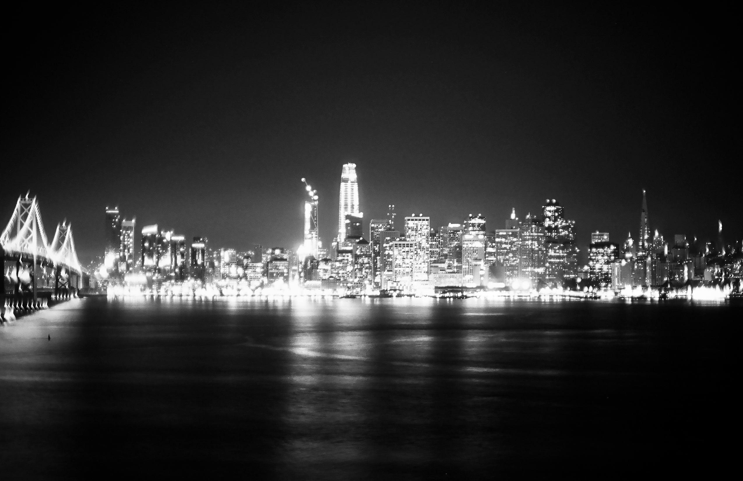 Panoramic nighttime skyline view showing illuminated Bay Bridge with suspension cables on left, glowing downtown buildings, and bay waters in foreground