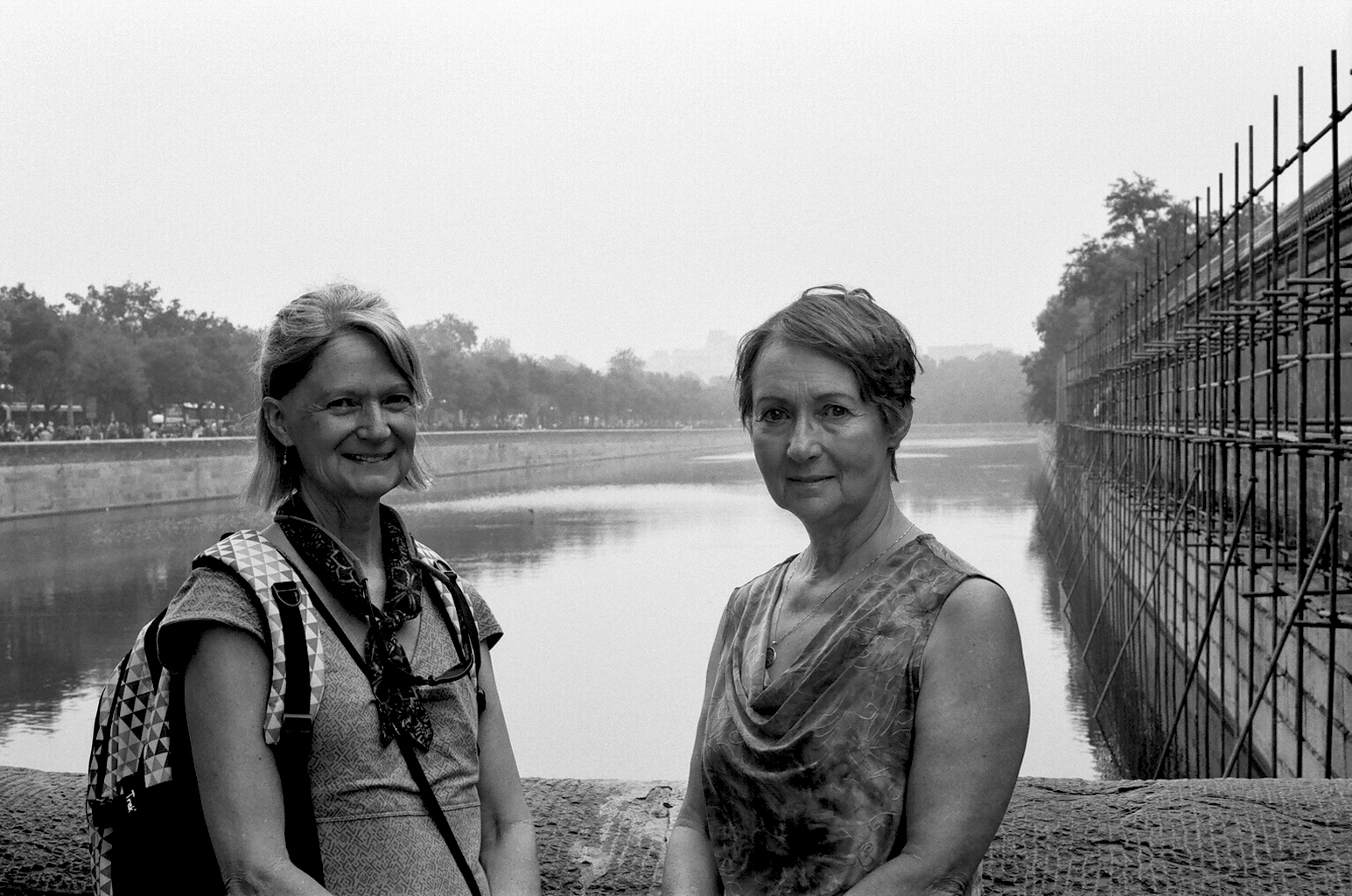 Two women posing by water with scaffolding and fortress wall in background