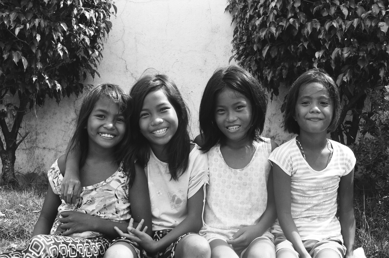 Four young Filipino girls sitting together smiling happily posed against wall with foliage