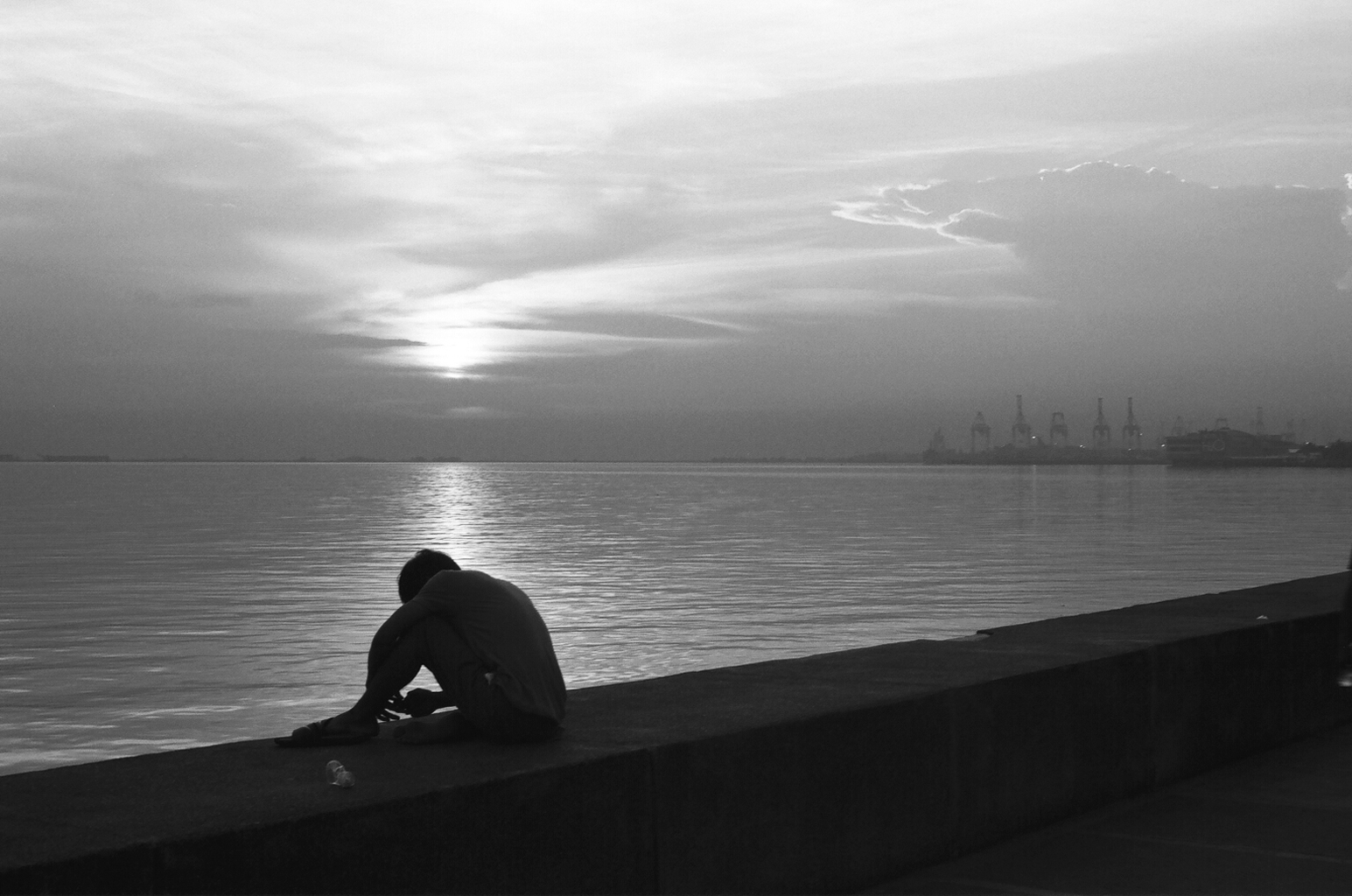 Person sitting alone on seawall at sunset with industrial harbor cranes visible in distance