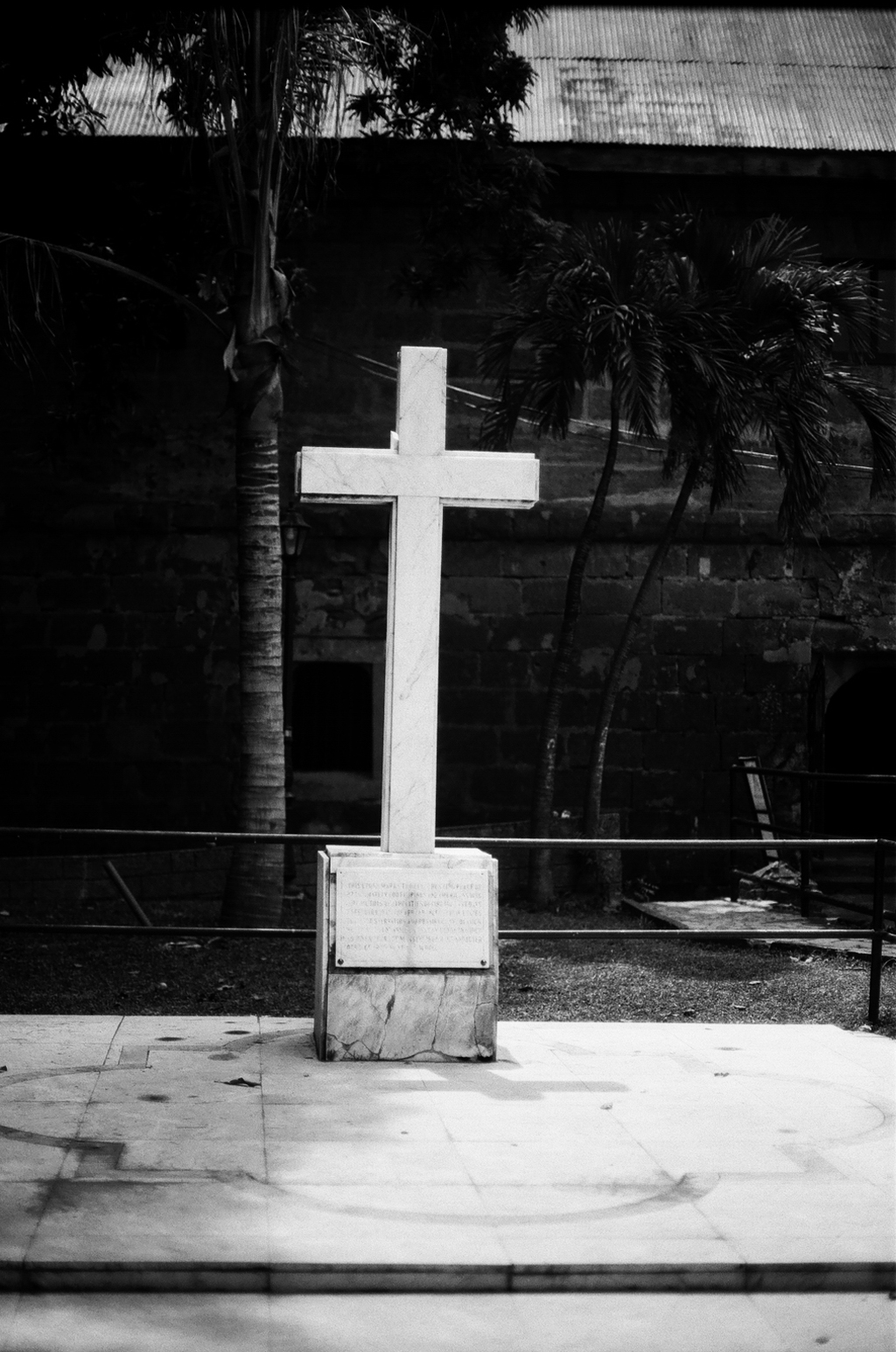 White stone cross memorial monument in courtyard with palm trees and old stone building
