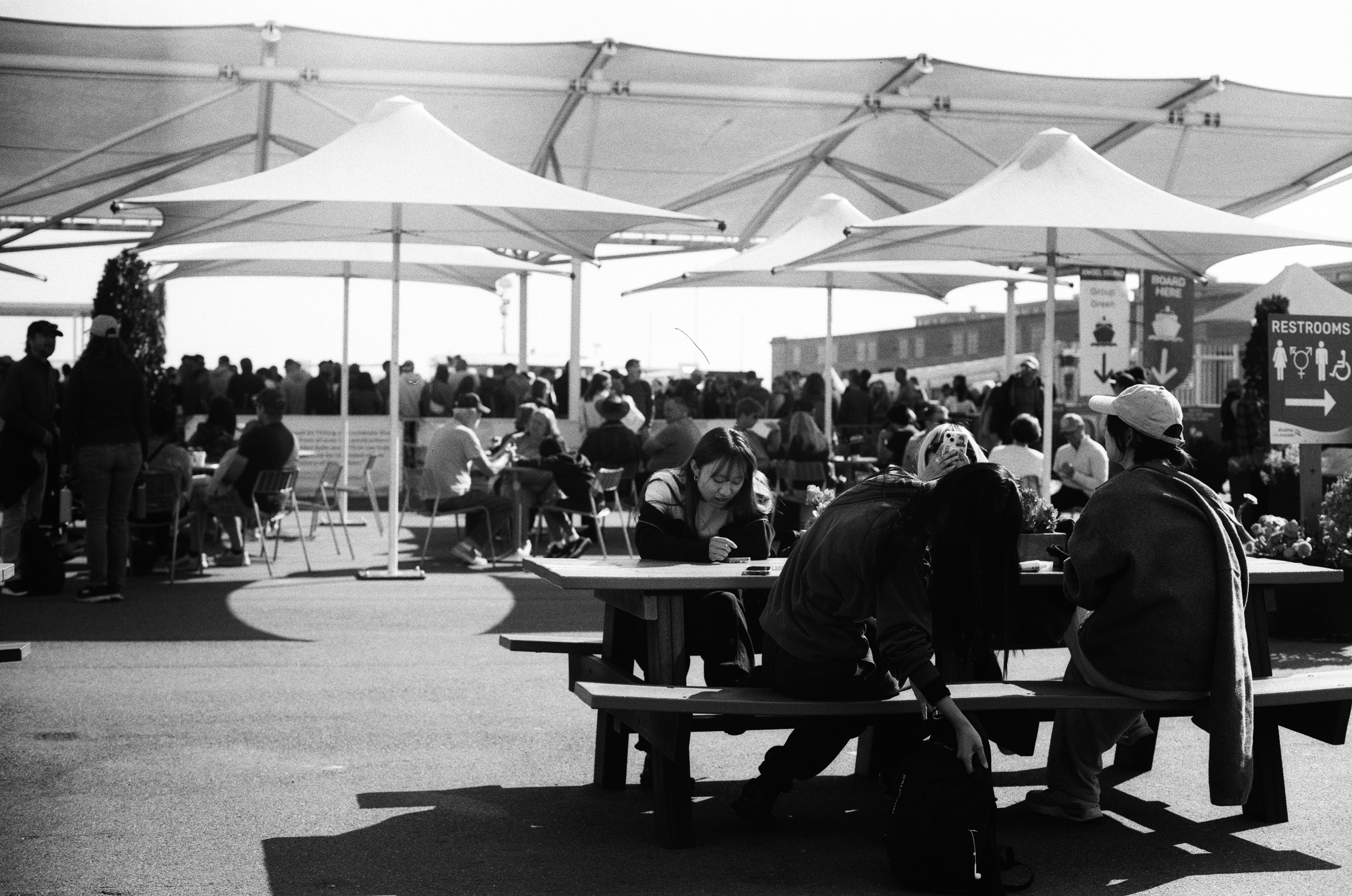 Waterfront marketplace with white shade sails and crowds of people with bay behind