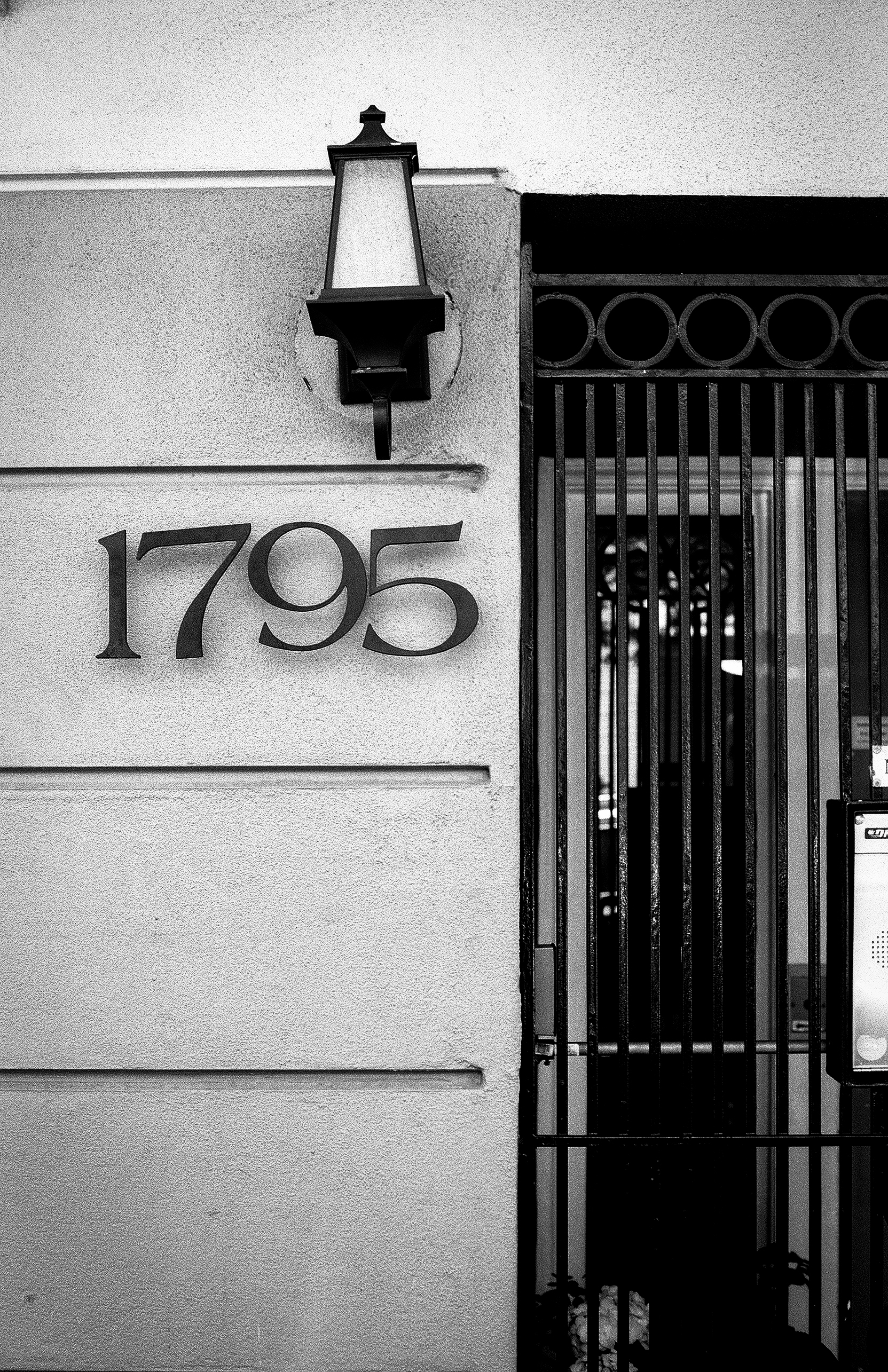 Address numbers 1795 on light concrete wall with sconce fixture and dark ornamental iron gate featuring geometric patterns