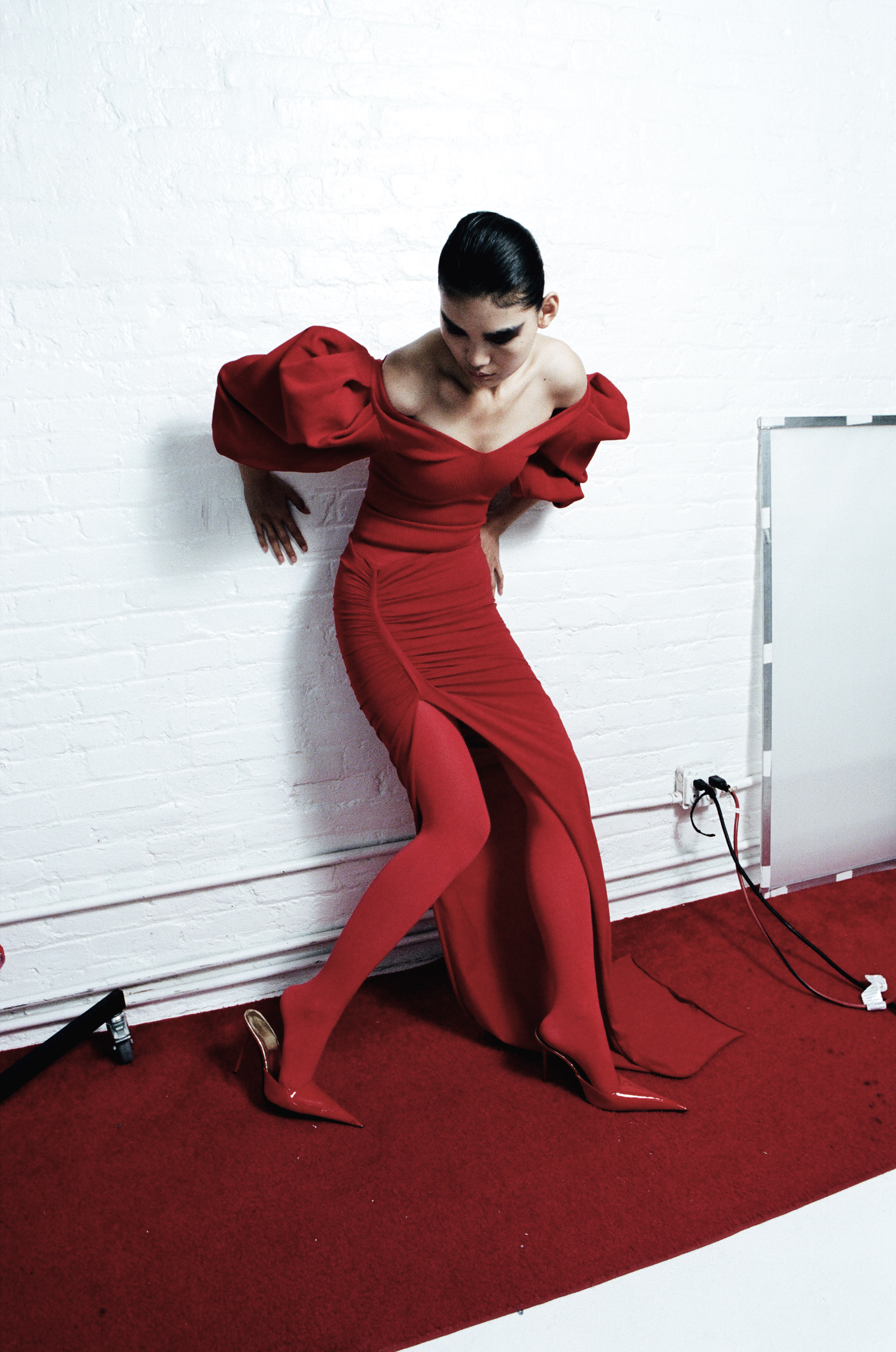 Model in dramatic red puff-sleeve gown with high slit against white brick wall