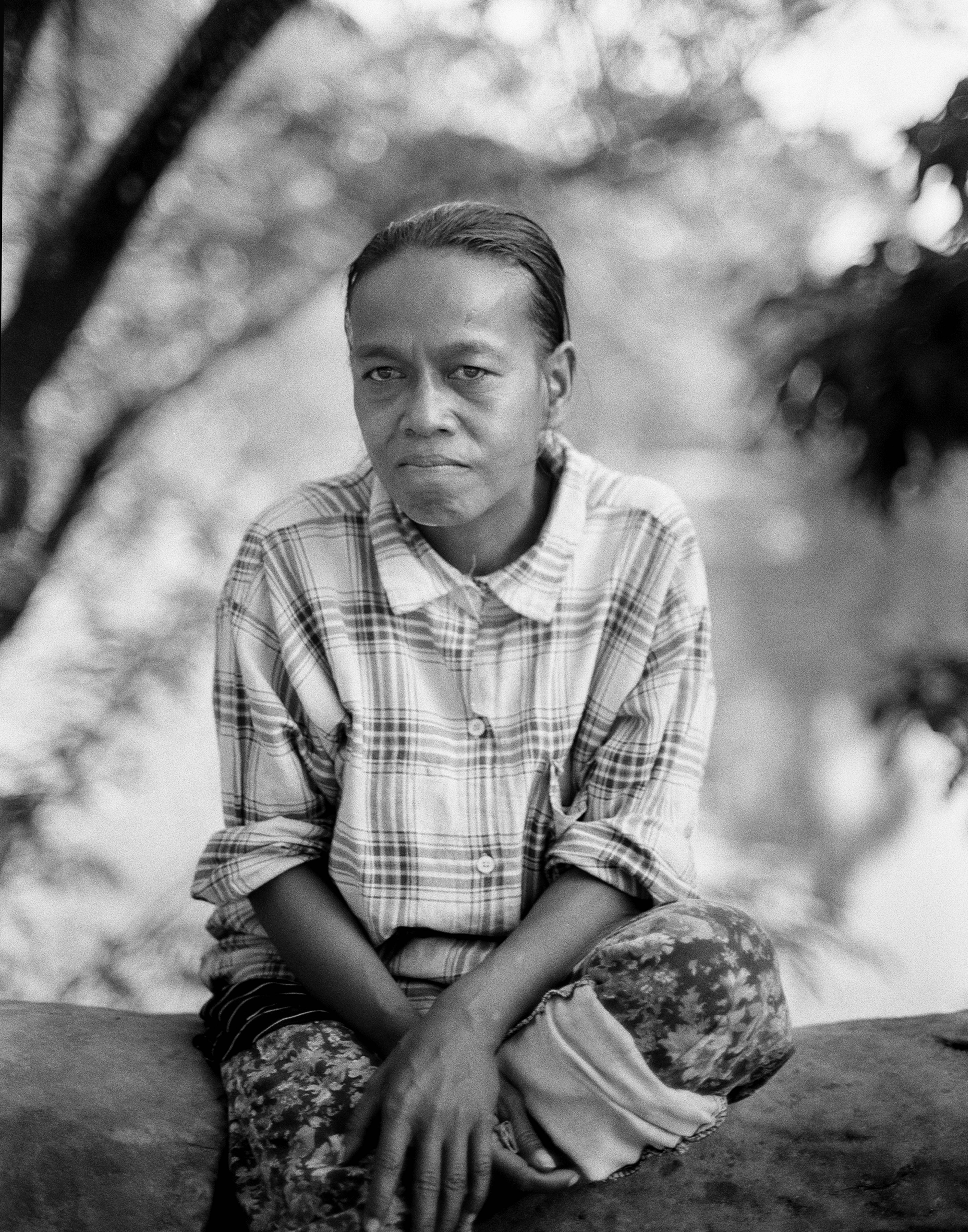Portrait of mature Cambodian woman in plaid shirt seated outdoors with weathered expression