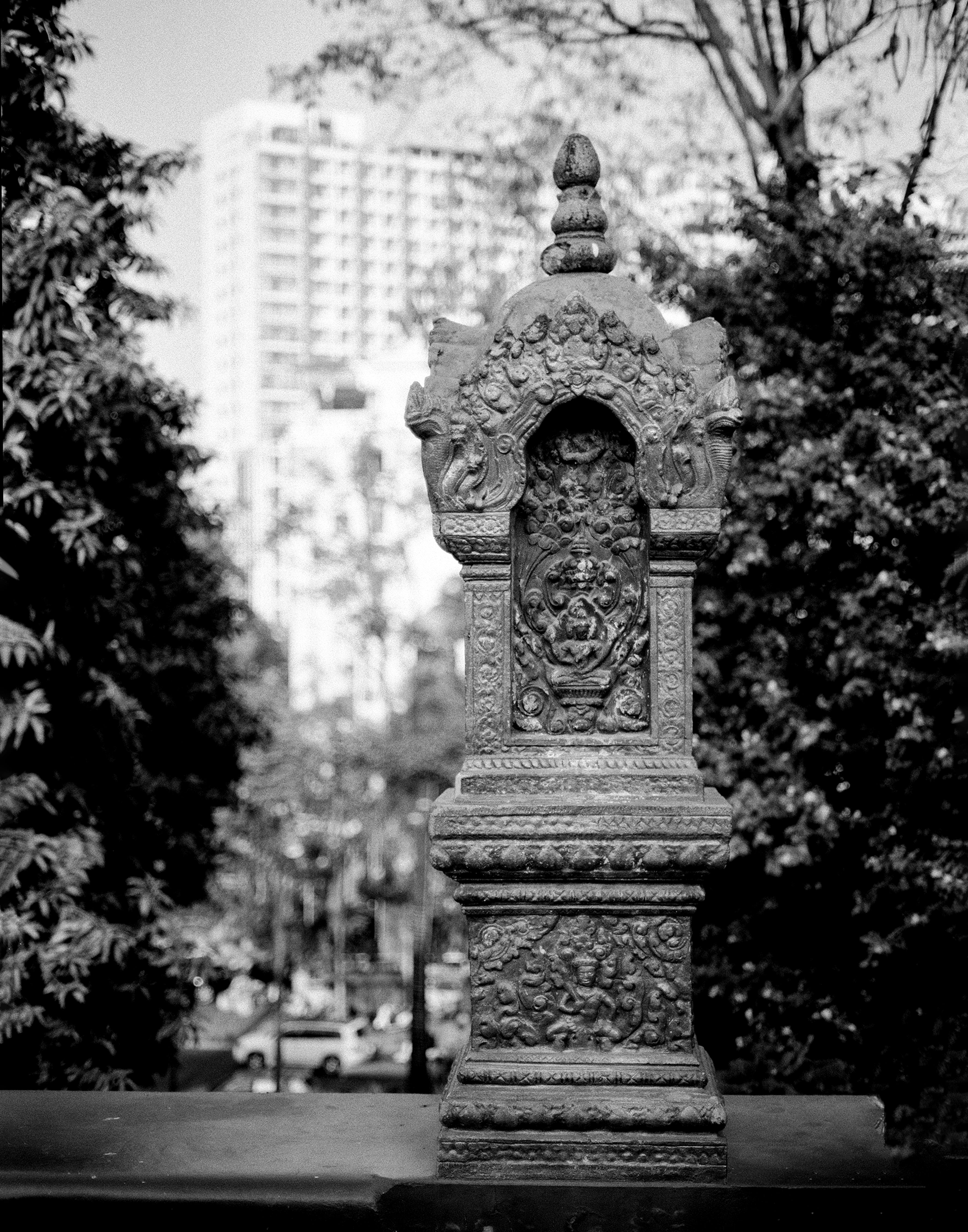 Ornate stone pedestal with carved demon guardian face framed by tropical foliage and modern buildings