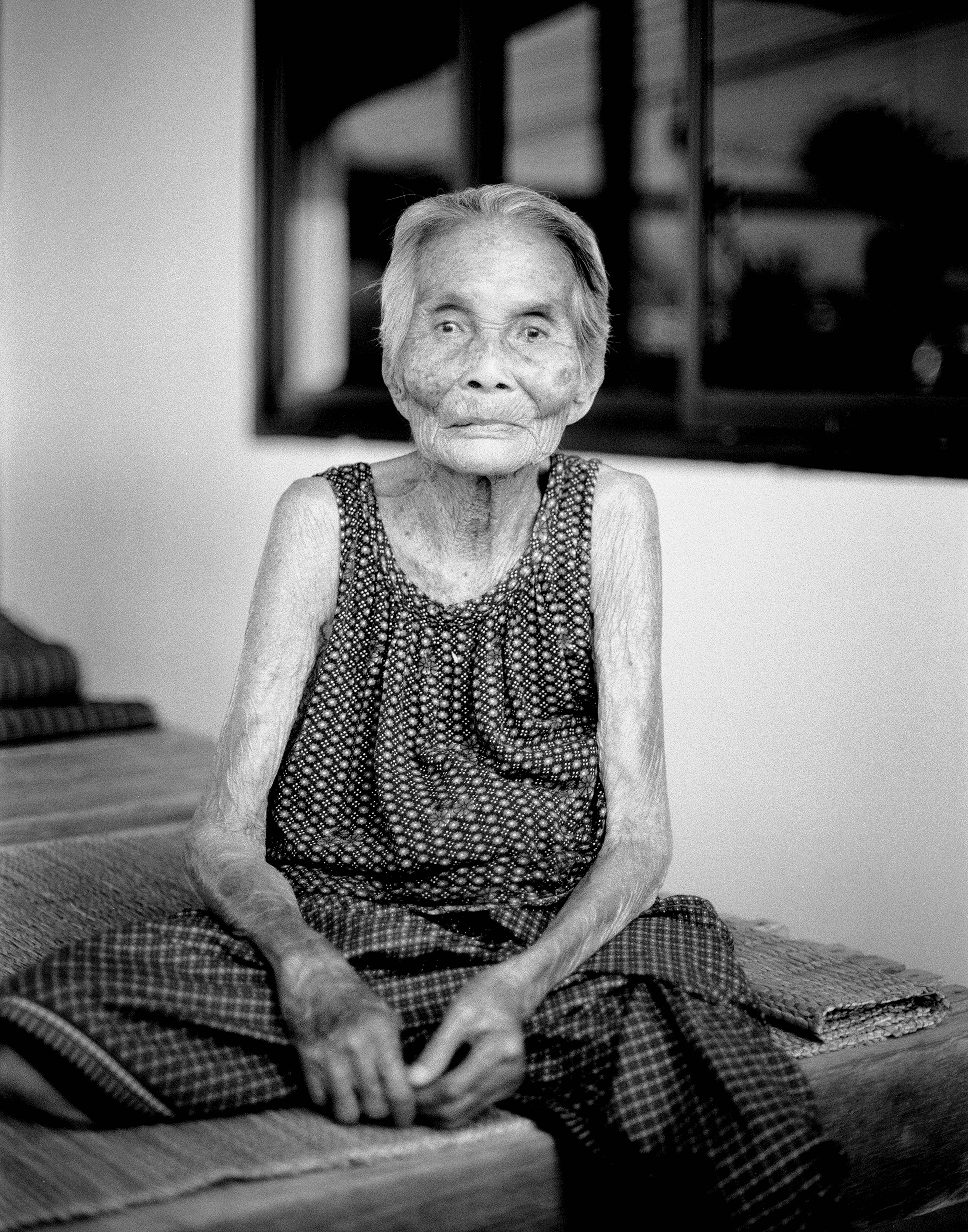 Elderly Thai woman in patterned dress seated on woven mat with dignified expression and weathered hands