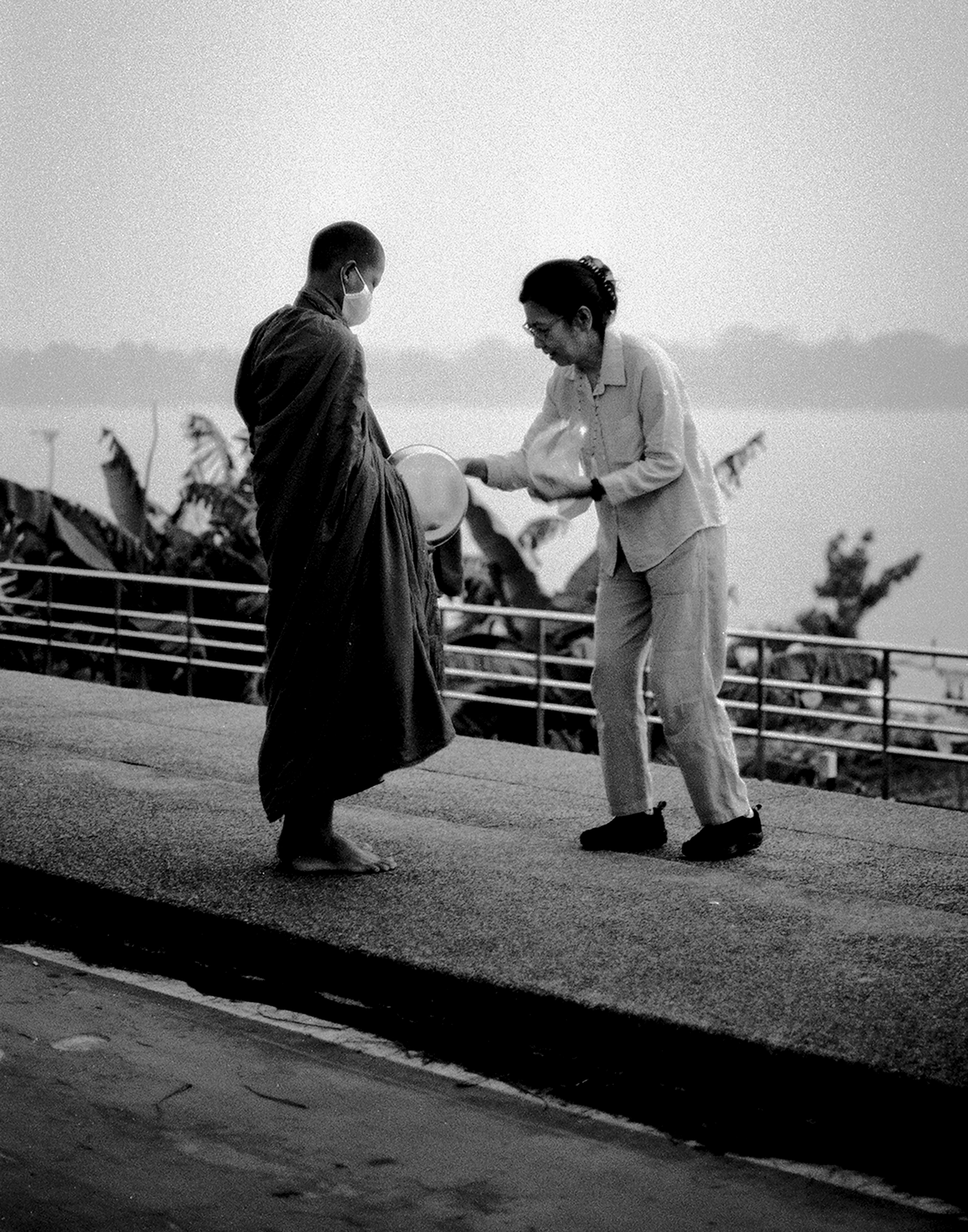 Buddhist monk receiving morning alms from woman on riverside promenade at dawn with misty Mekong background