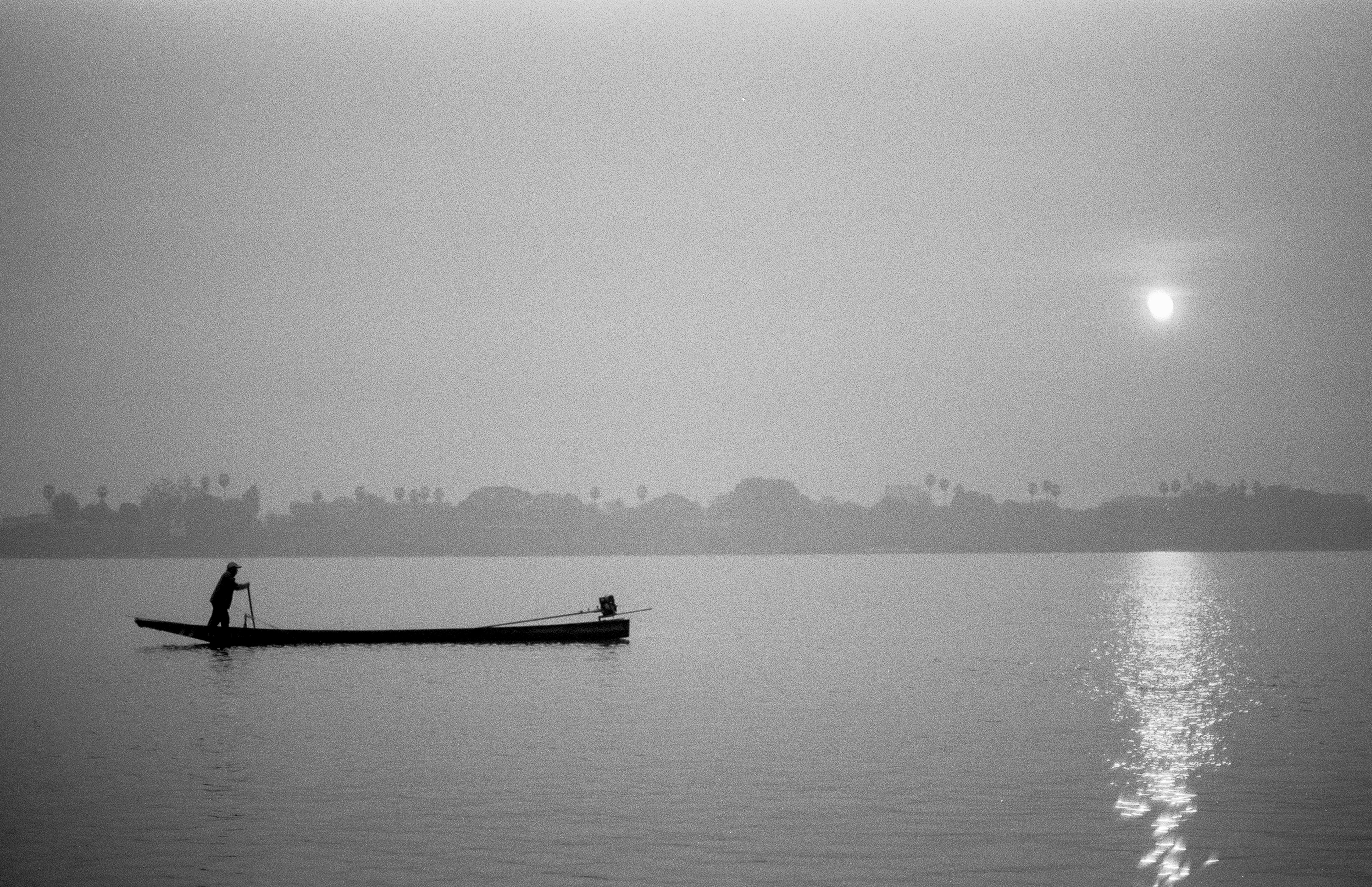 Lone fisherman on long-tail boat silhouetted on Mekong River at dawn with sun reflection on water