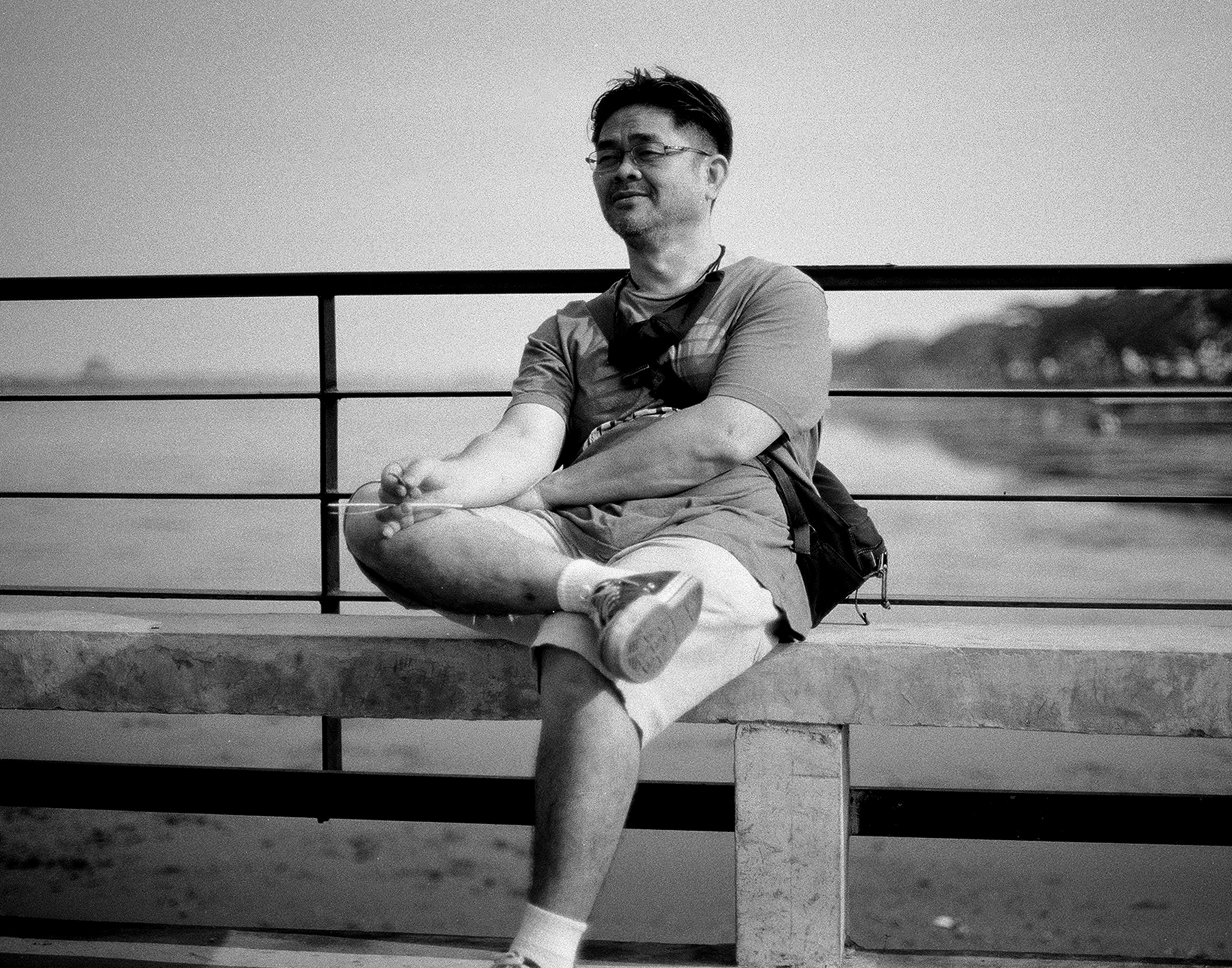 Middle-aged man with glasses relaxing on concrete bench by riverside in casual contemplative pose
