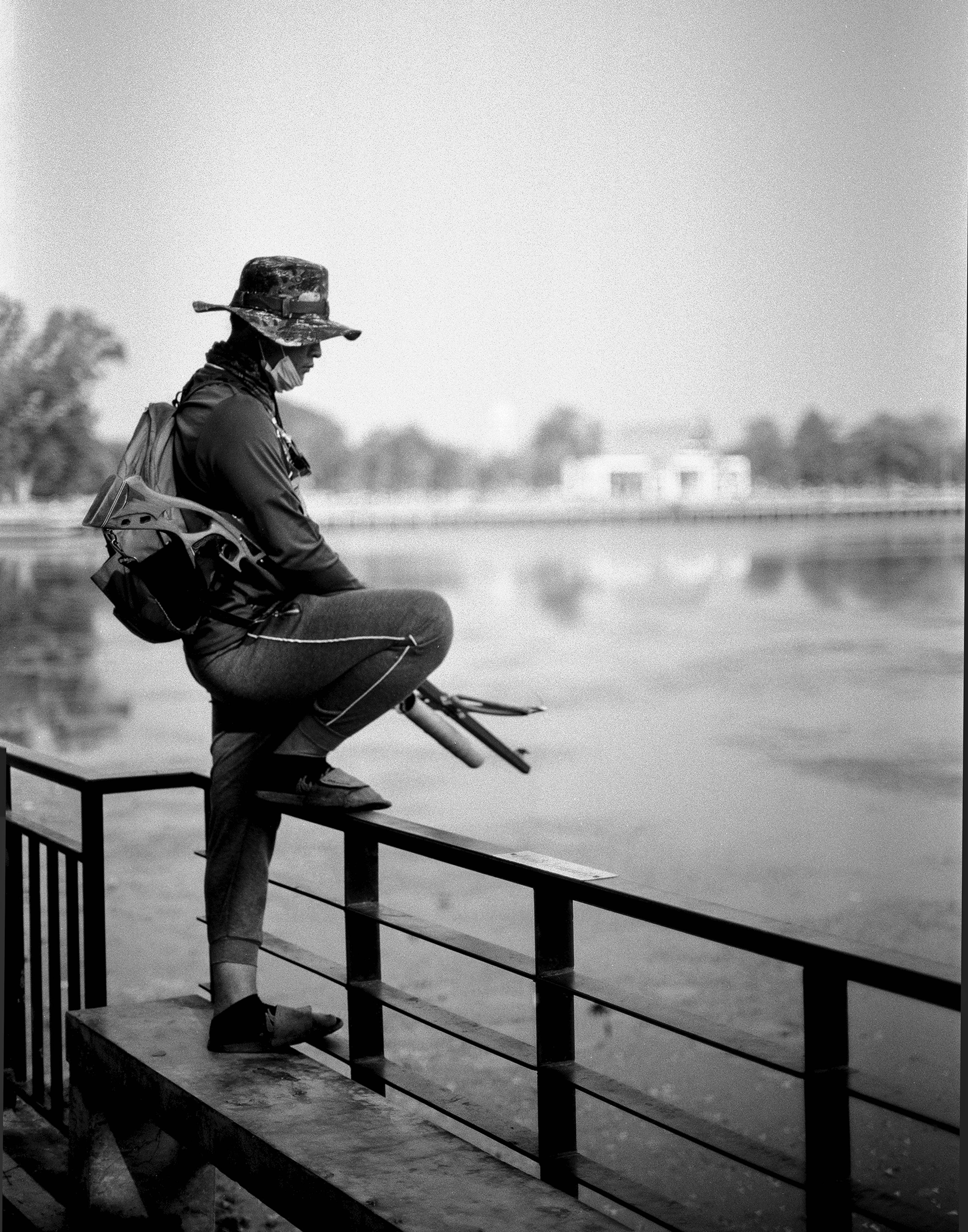 Person with hat and face mask crouching on railing by calm river photographing the serene waterscape