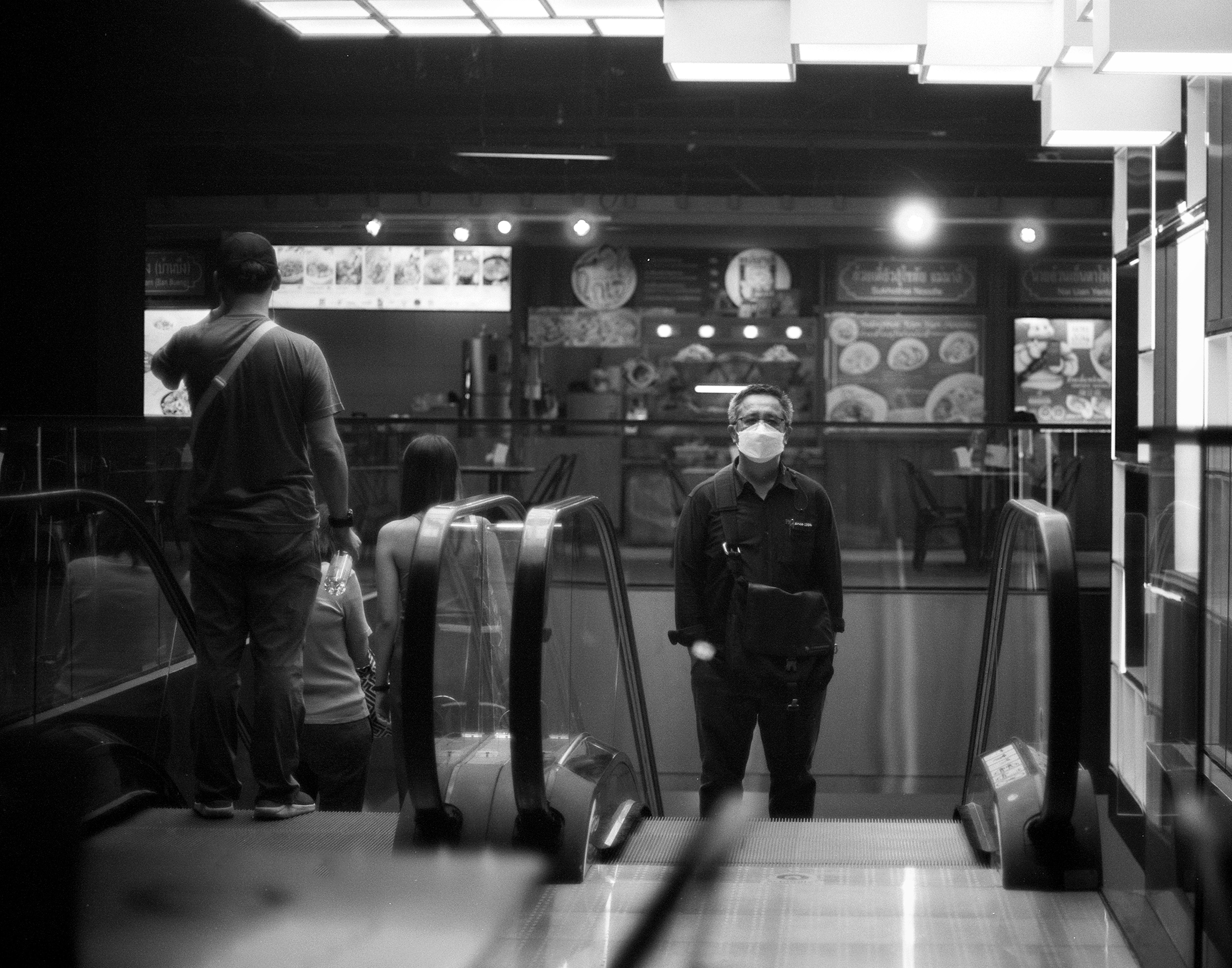 Man wearing face mask standing between escalators in shopping mall food court with Thai signage and illuminated menu boards