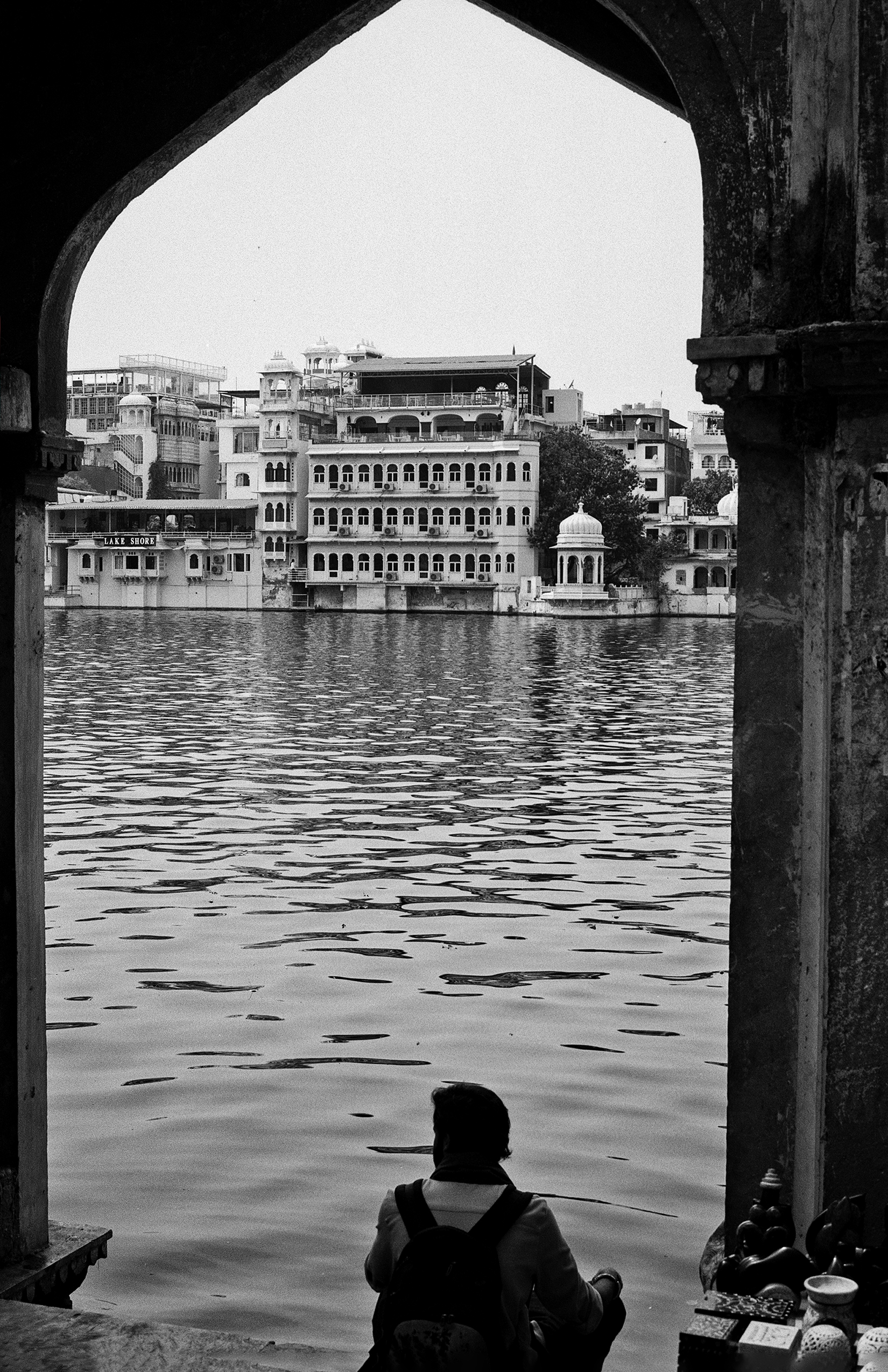 Black and white person seated at archway viewing Udaipur lake palace across water ornate architecture