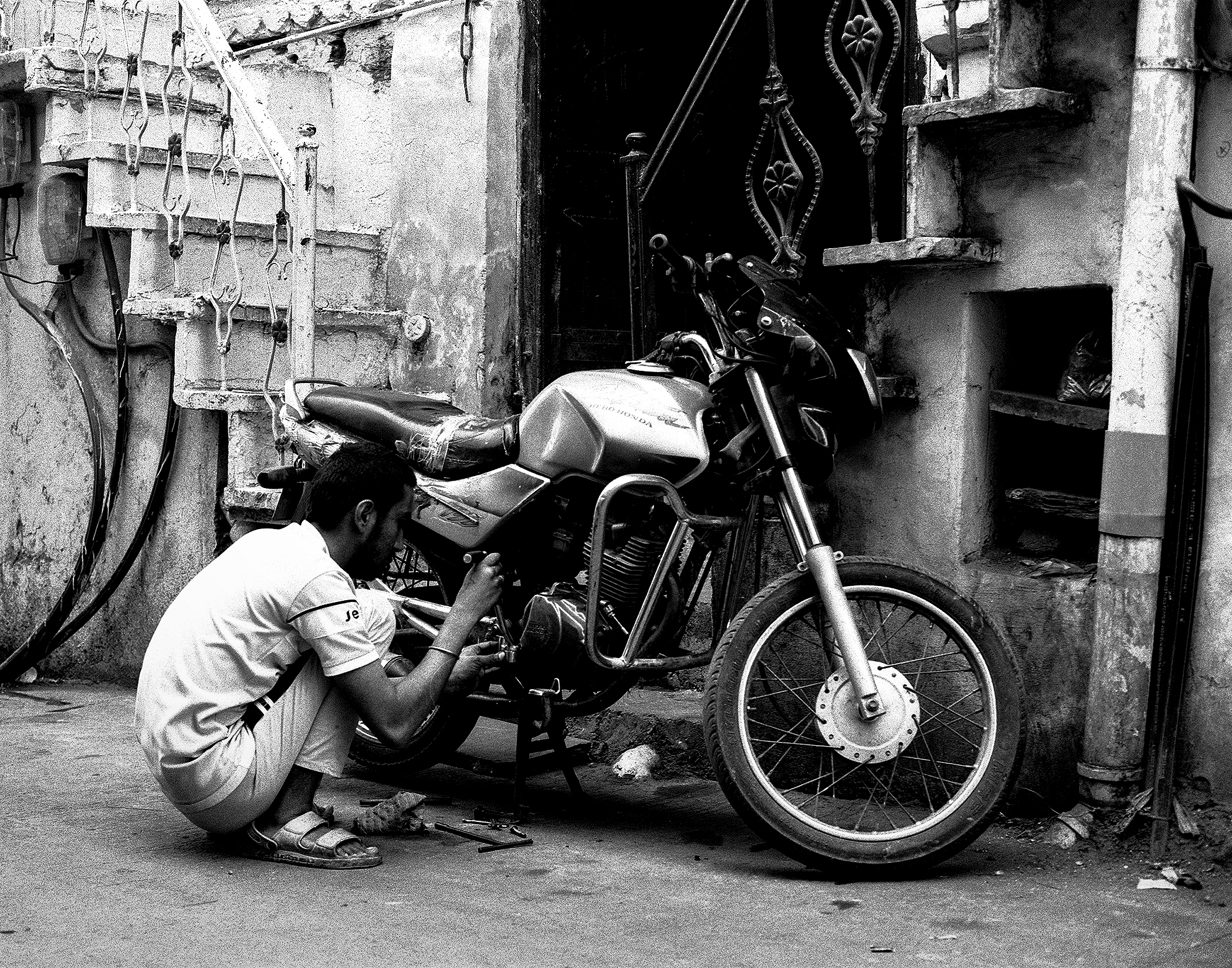 Black and white mechanic crouching beside motorcycle working on engine urban alley setting
