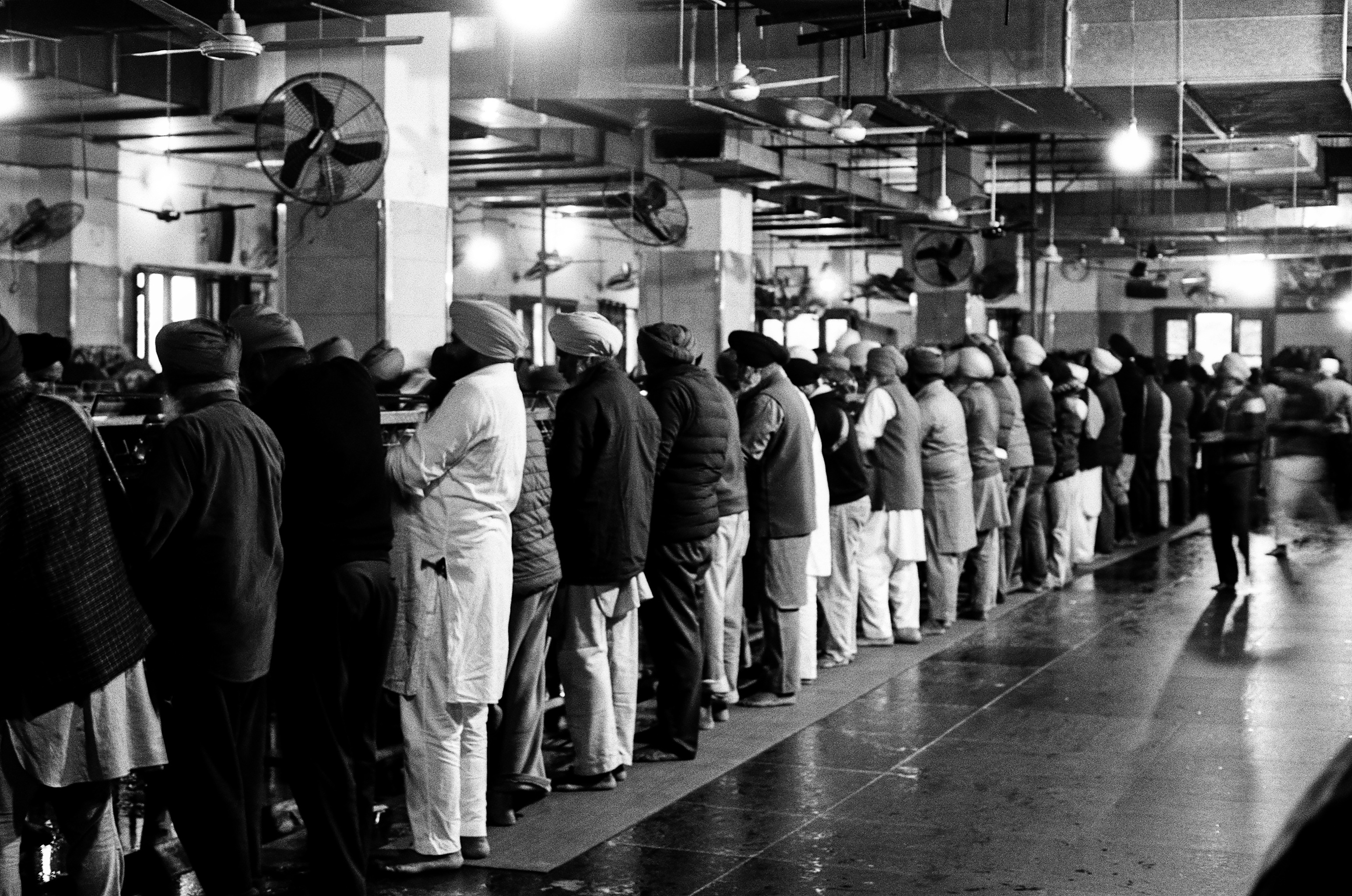Black and white ceremonial crowd of men in traditional dress standing in factory-like industrial hall with fans