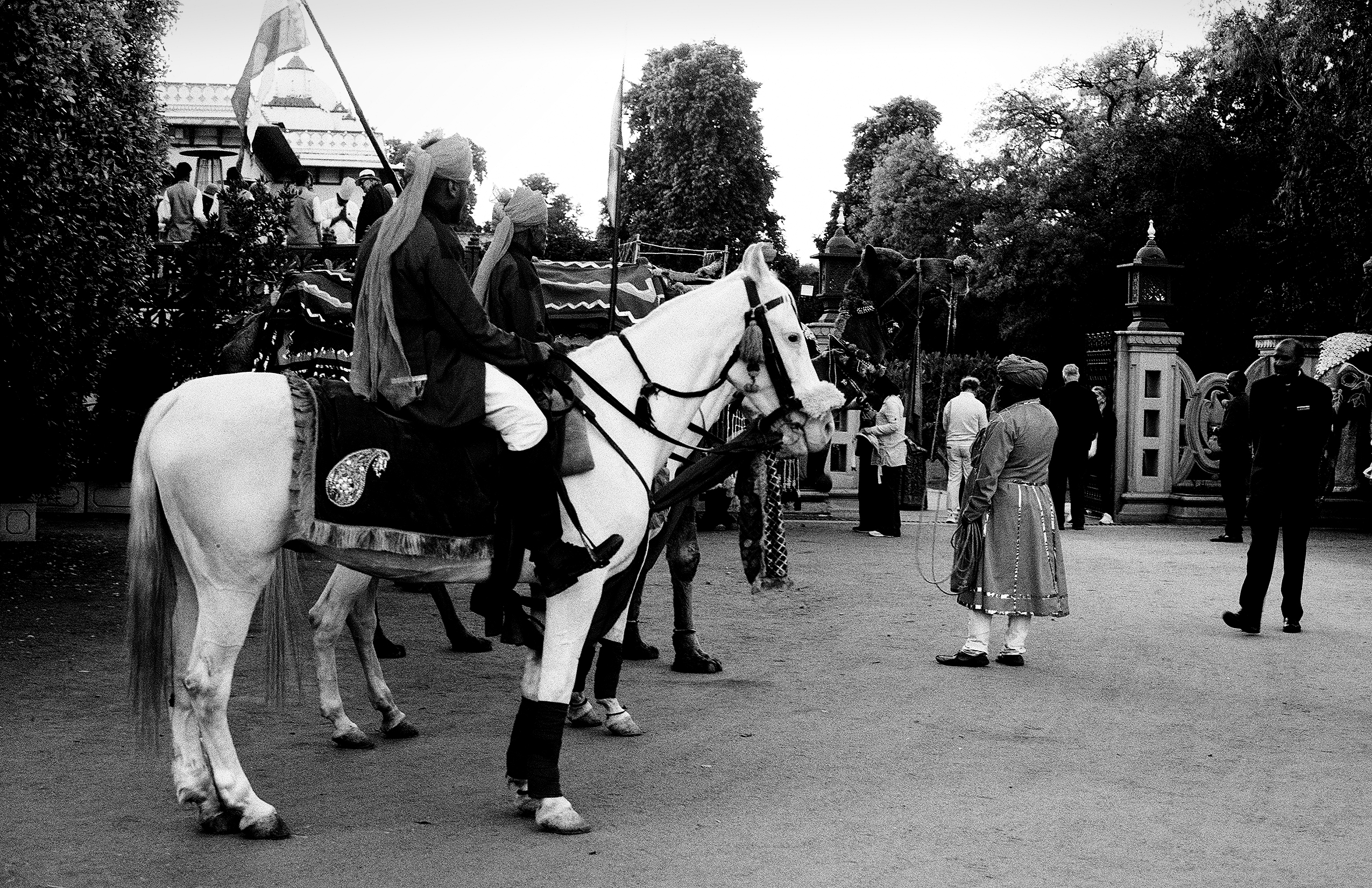 Black and white maharaja in ceremonial robes on white horse in front of palace with attendants and crowds