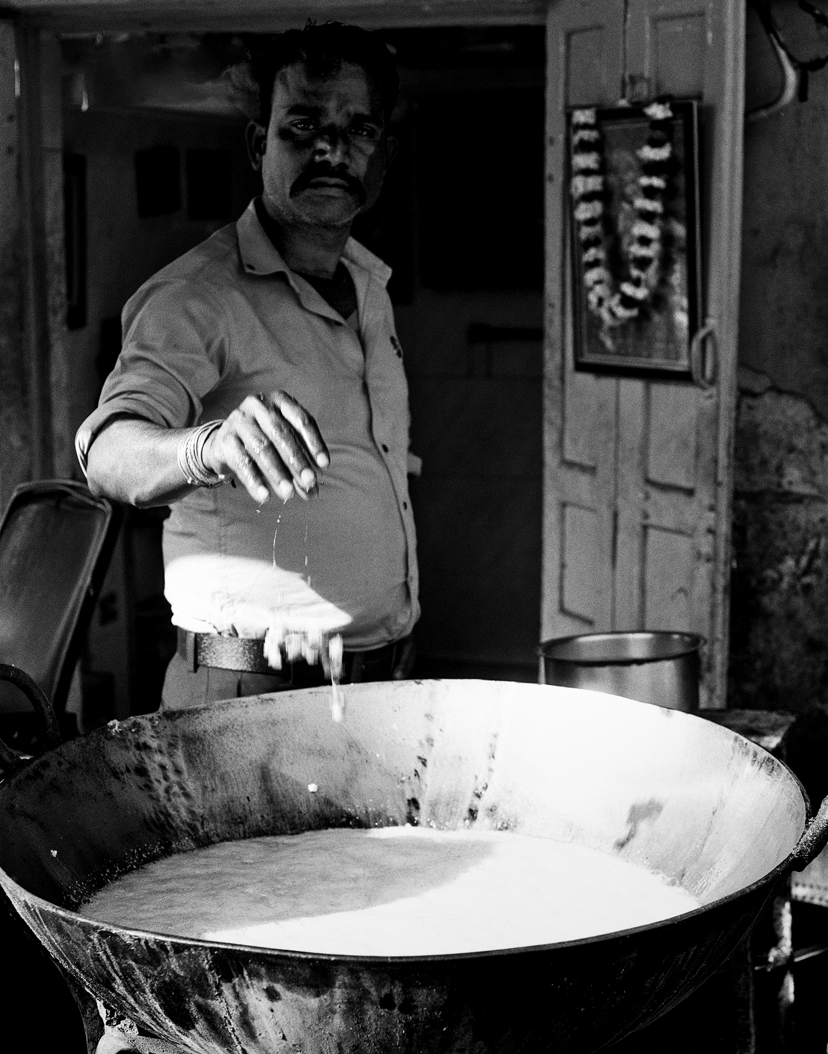 Black and white man in light colored shirt preparing traditional Indian sweets pouring mixture in metal pan