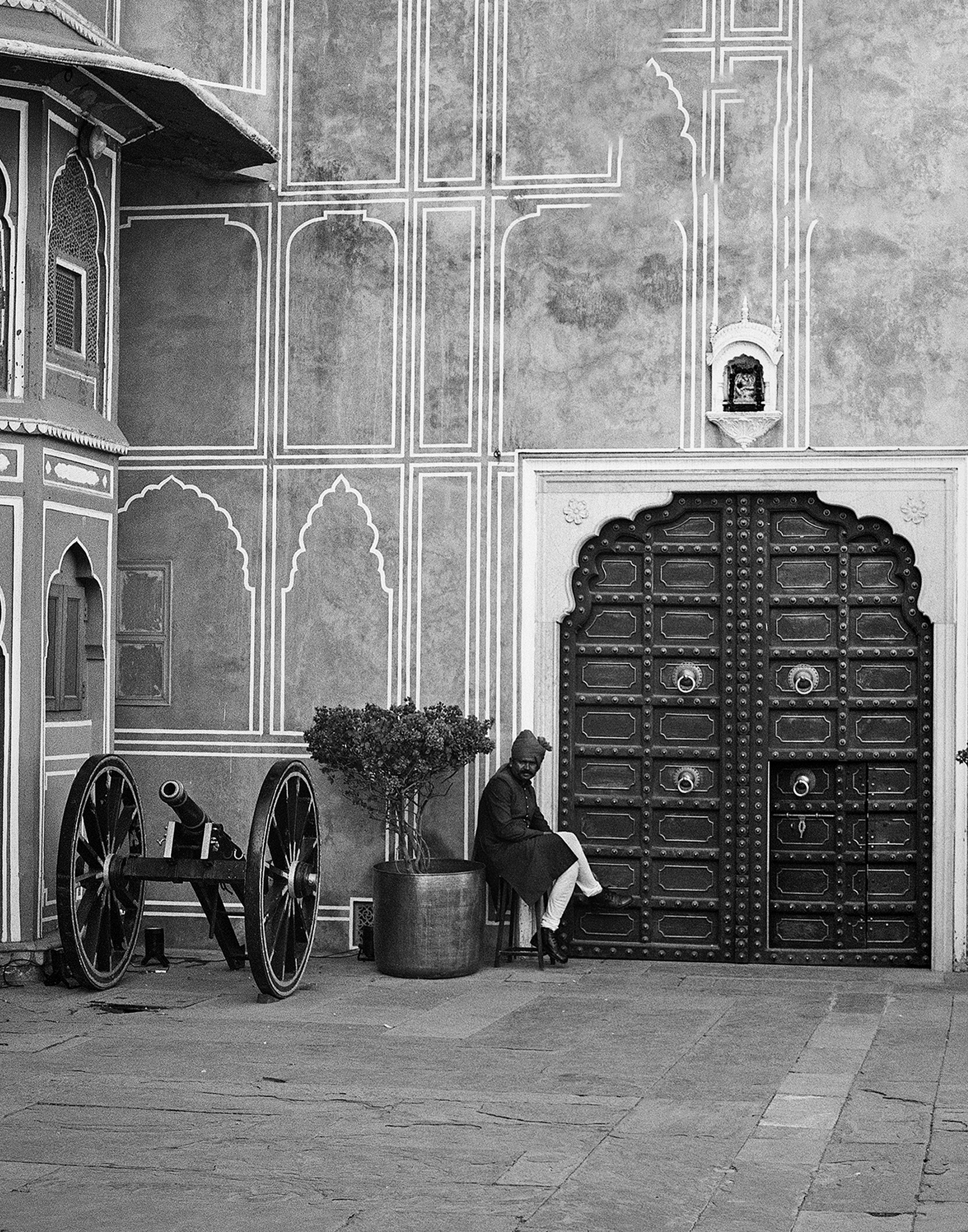 Black and white man sitting before palace wall with ornate arches decorative door and cannon display