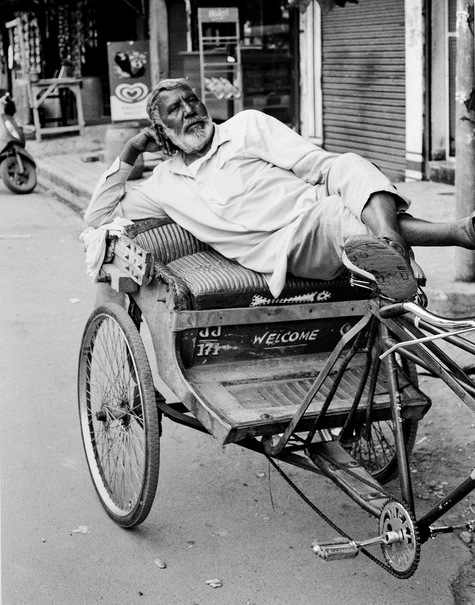 Black and white elderly man resting reclined in hand-pulled cart labeled welcome on urban street
