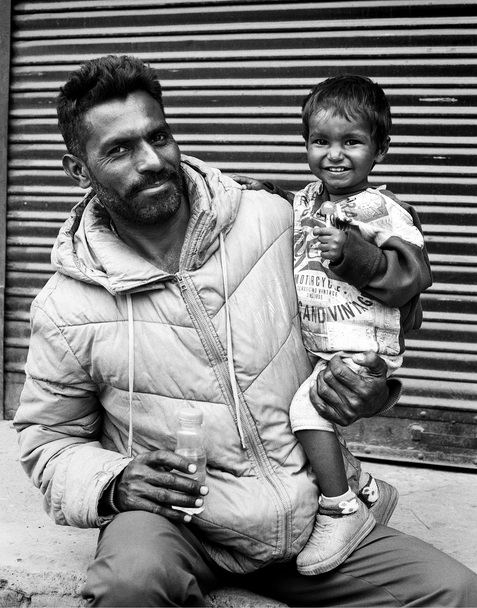 Black and white portrait father holding young smiling child wearing winter jacket against metal shutter