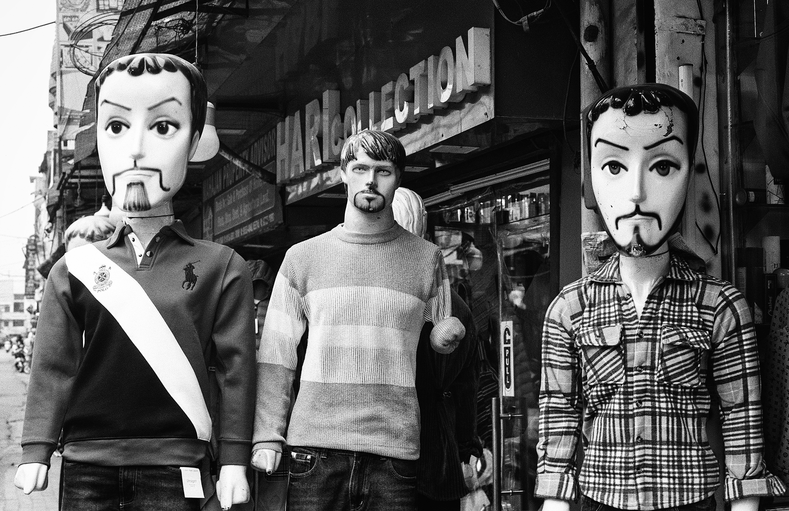 Black and white surreal street scene with three mannequin figures wearing elaborate masks clothing