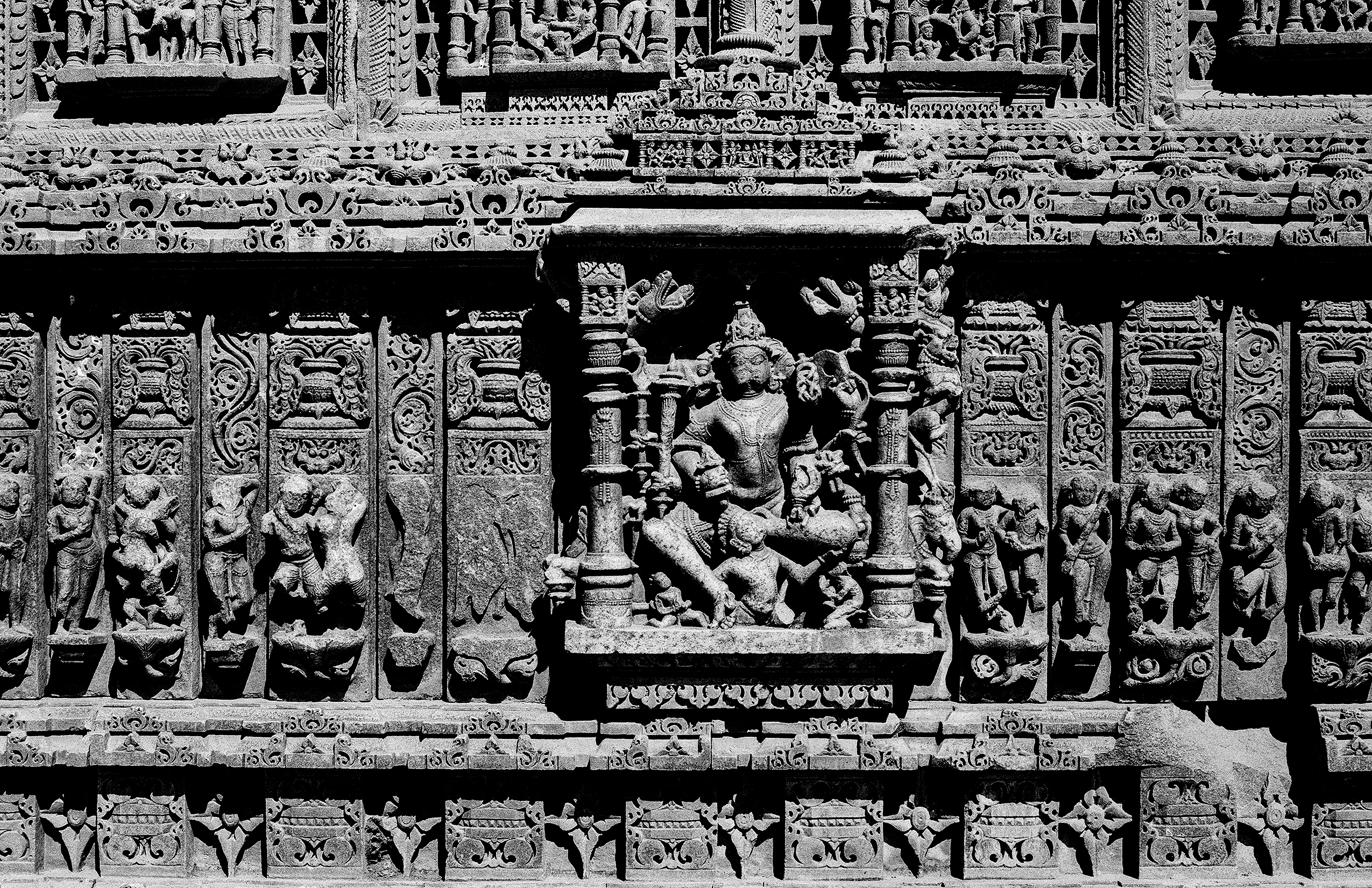 Black and white intricate temple wall relief carving showing multiple deity figures and decorative bands