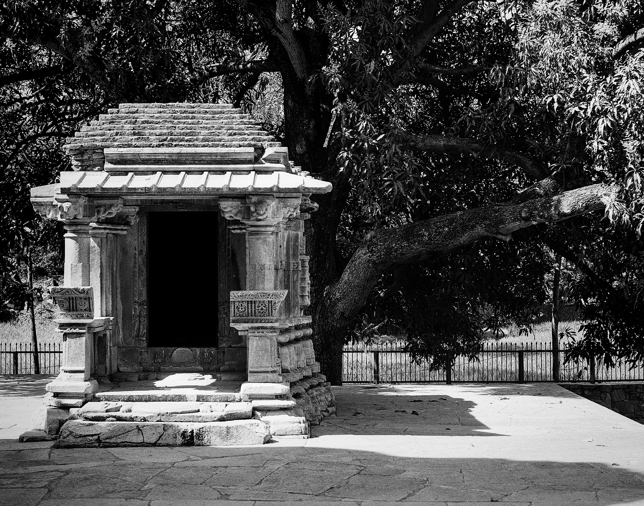 Black and white small ancient stone temple structure with tiered roof under large tree