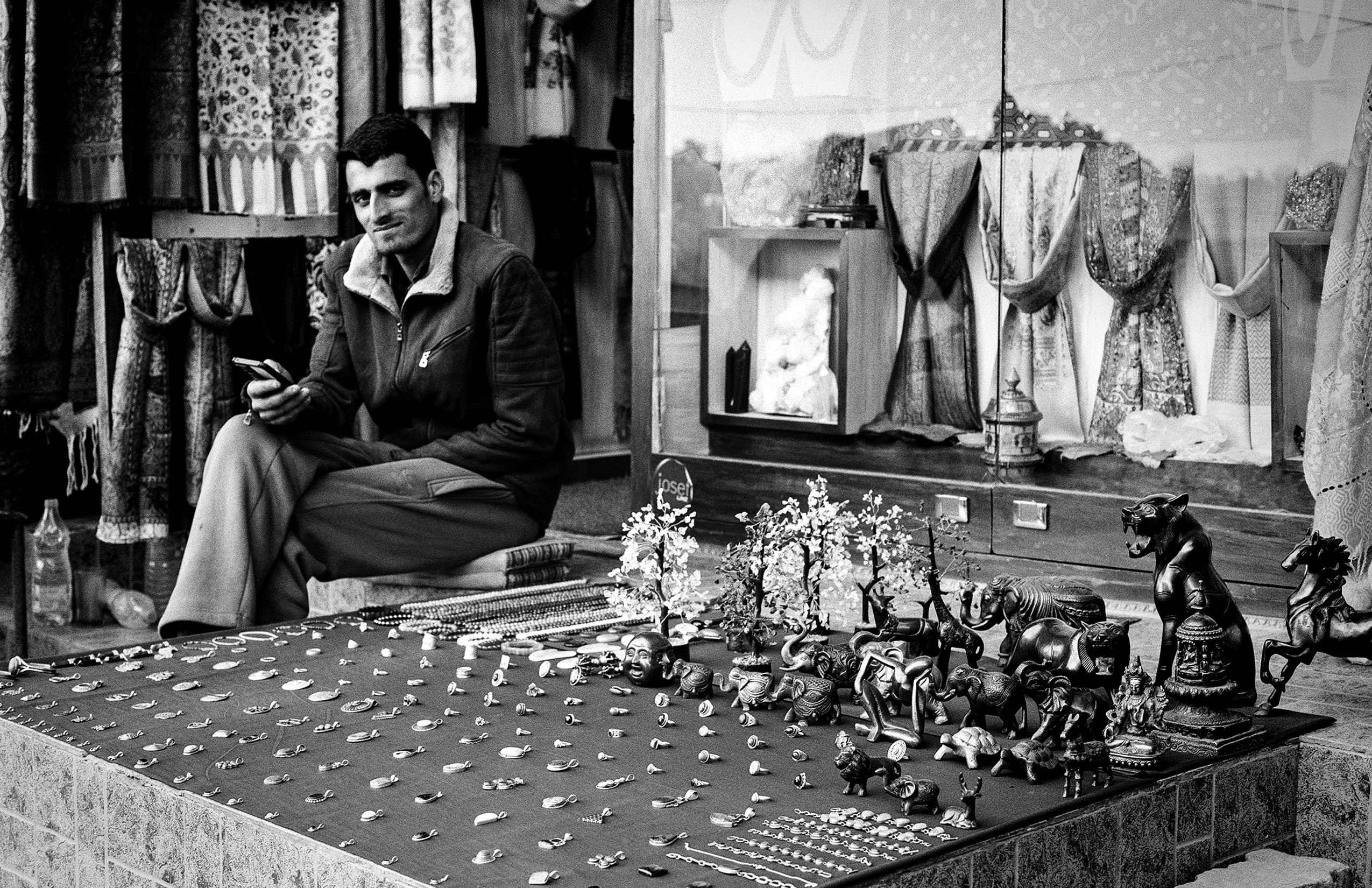 Market vendor sitting at stall with bronze figurines elephants jewelry and handicrafts colorful textiles hanging in background Dharamsala bazaar
