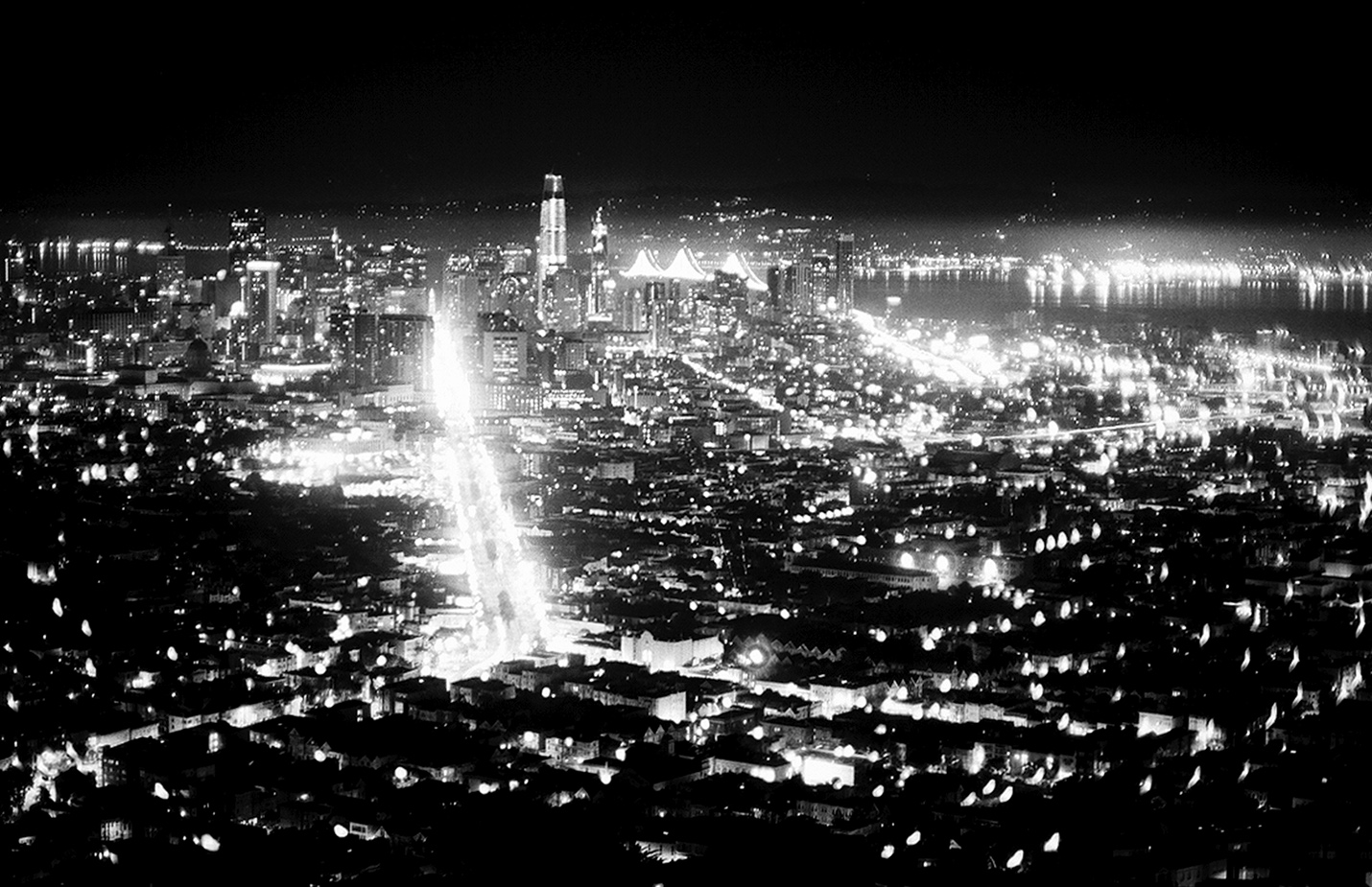 Panoramic nighttime cityscape with brilliant street lights creating vertical illumination, downtown skyline, bay area, and distant mountains