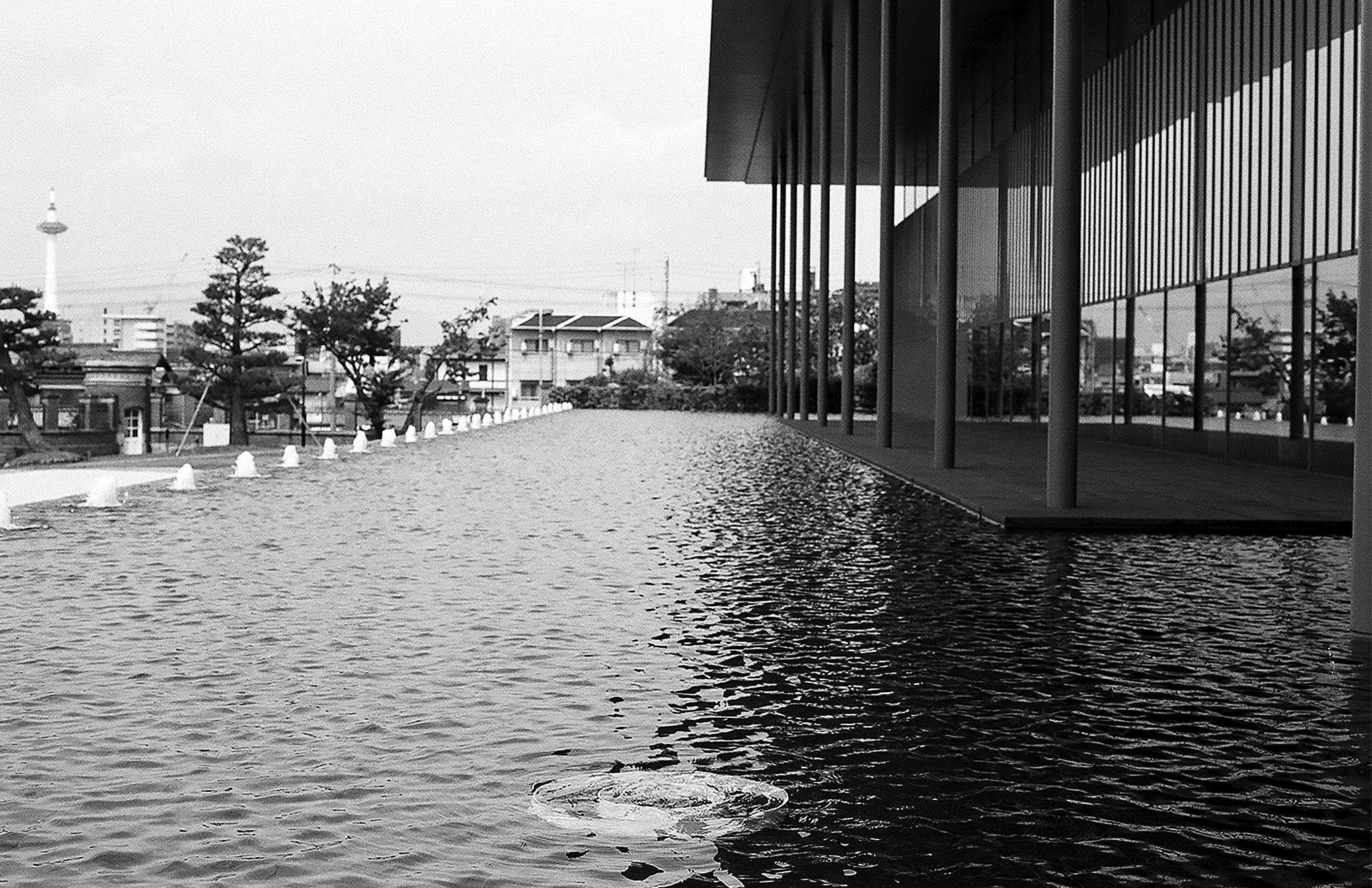 Contemporary pavilion with tall vertical post columns reflecting in still water with Japanese landscape distant buildings