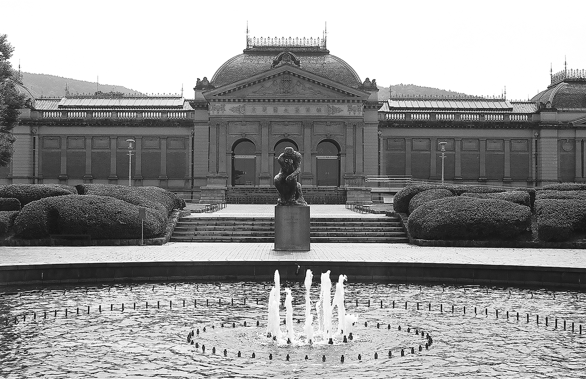 Grand fountain pond with neoclassical museum building Rodin Thinker sculpture and manicured hedges in plaza