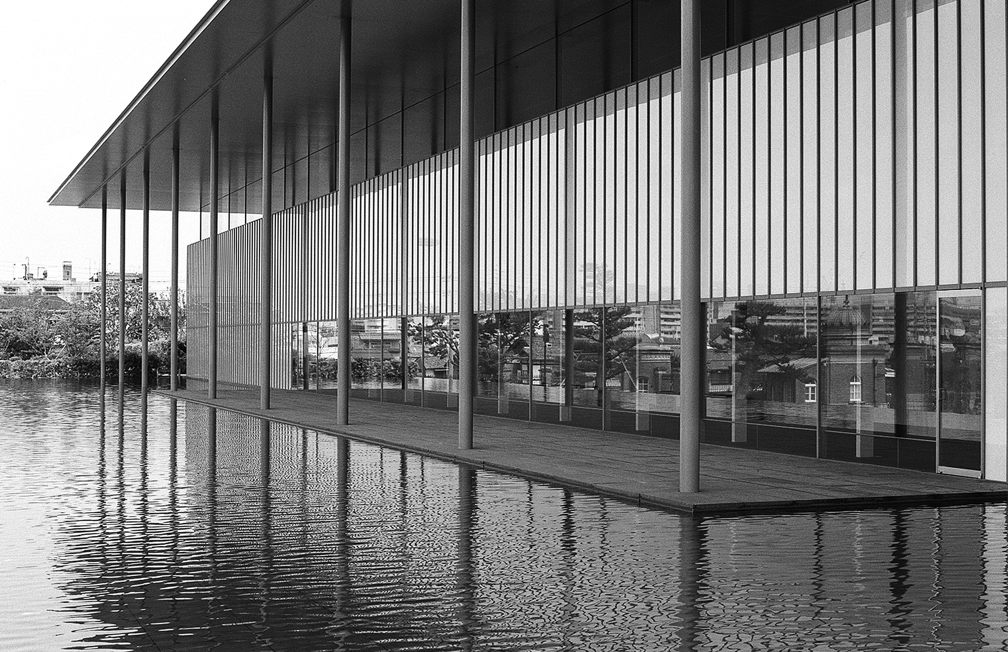 Long horizontal architectural view of modern pavilion with vertical columns reflecting in water pool and distant cityscape