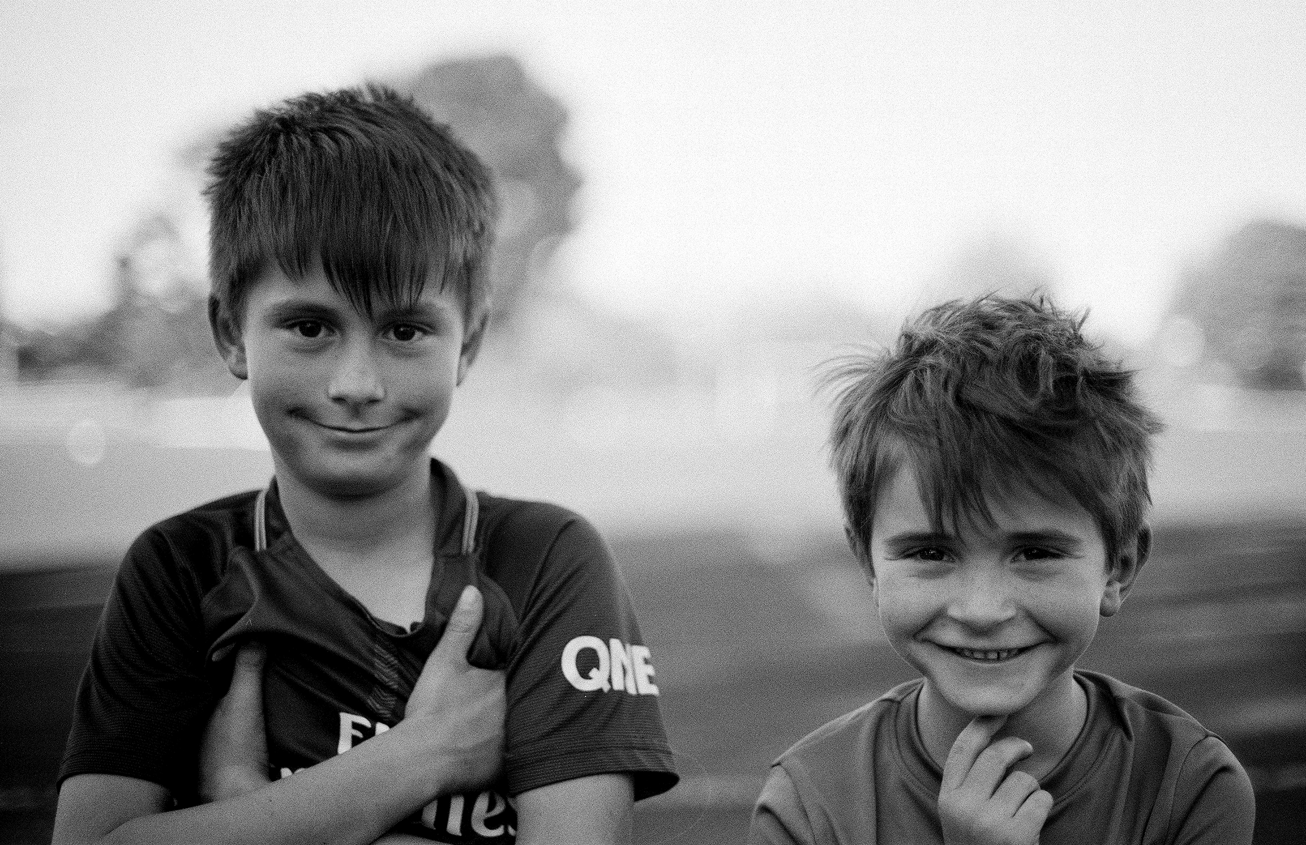 Two young boys smiling at camera one giving thumbs up outdoor portrait with soft bokeh background