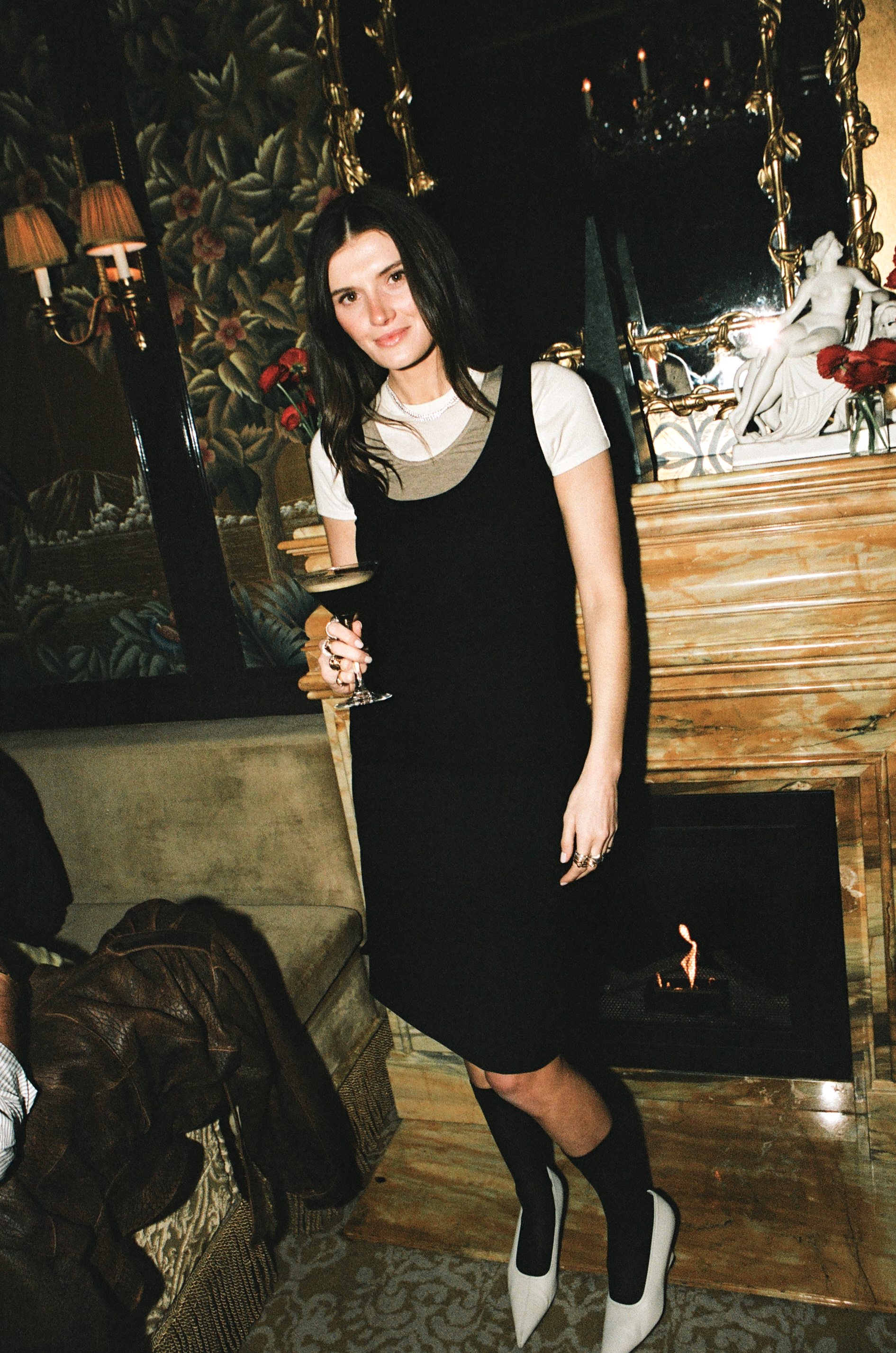 Woman in a black slip dress over a white tee with knee socks and white heels by a fireplace