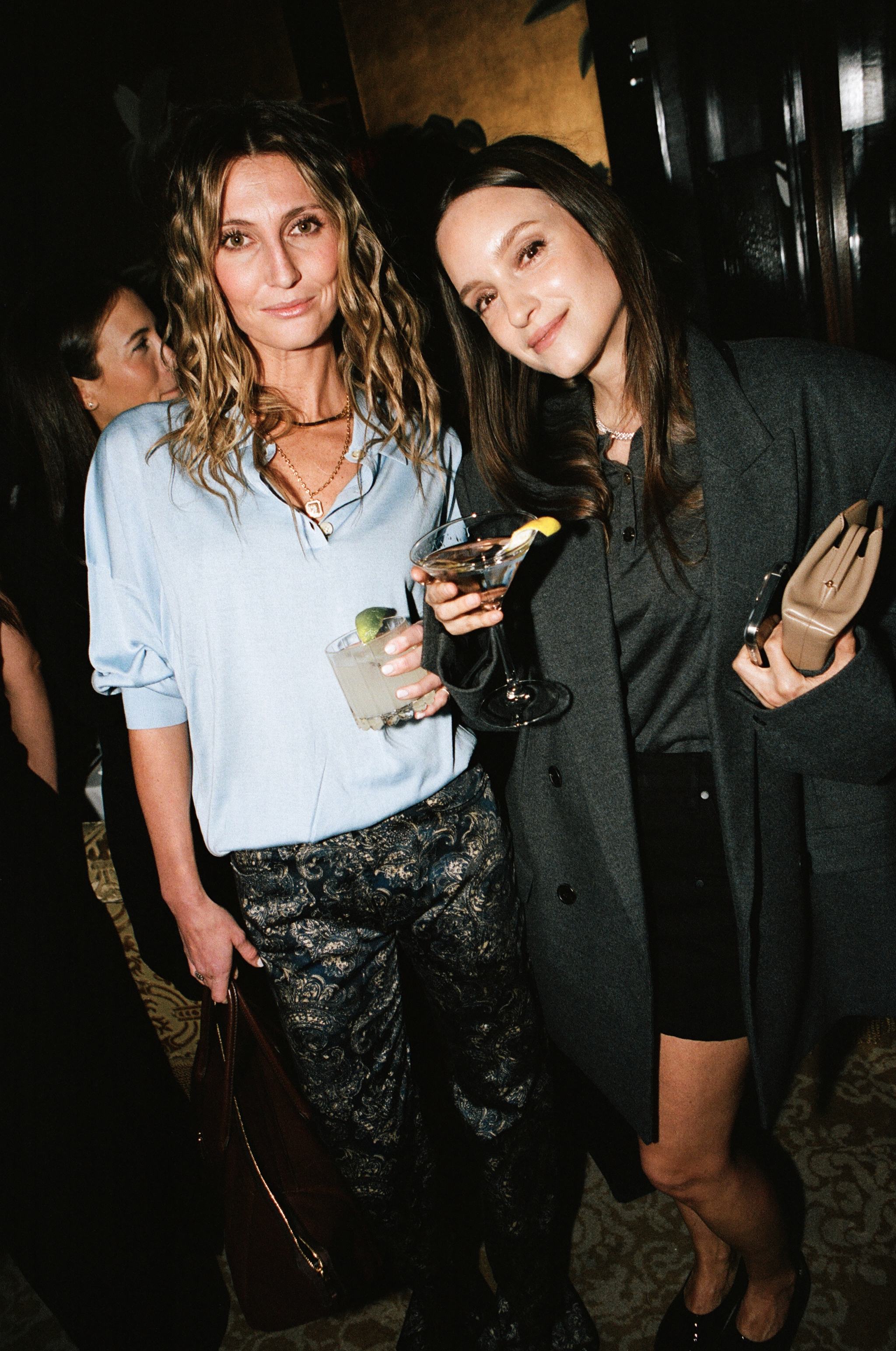 Two women at a party, one in a blue blouse with a margarita and one in a grey coat with a martini