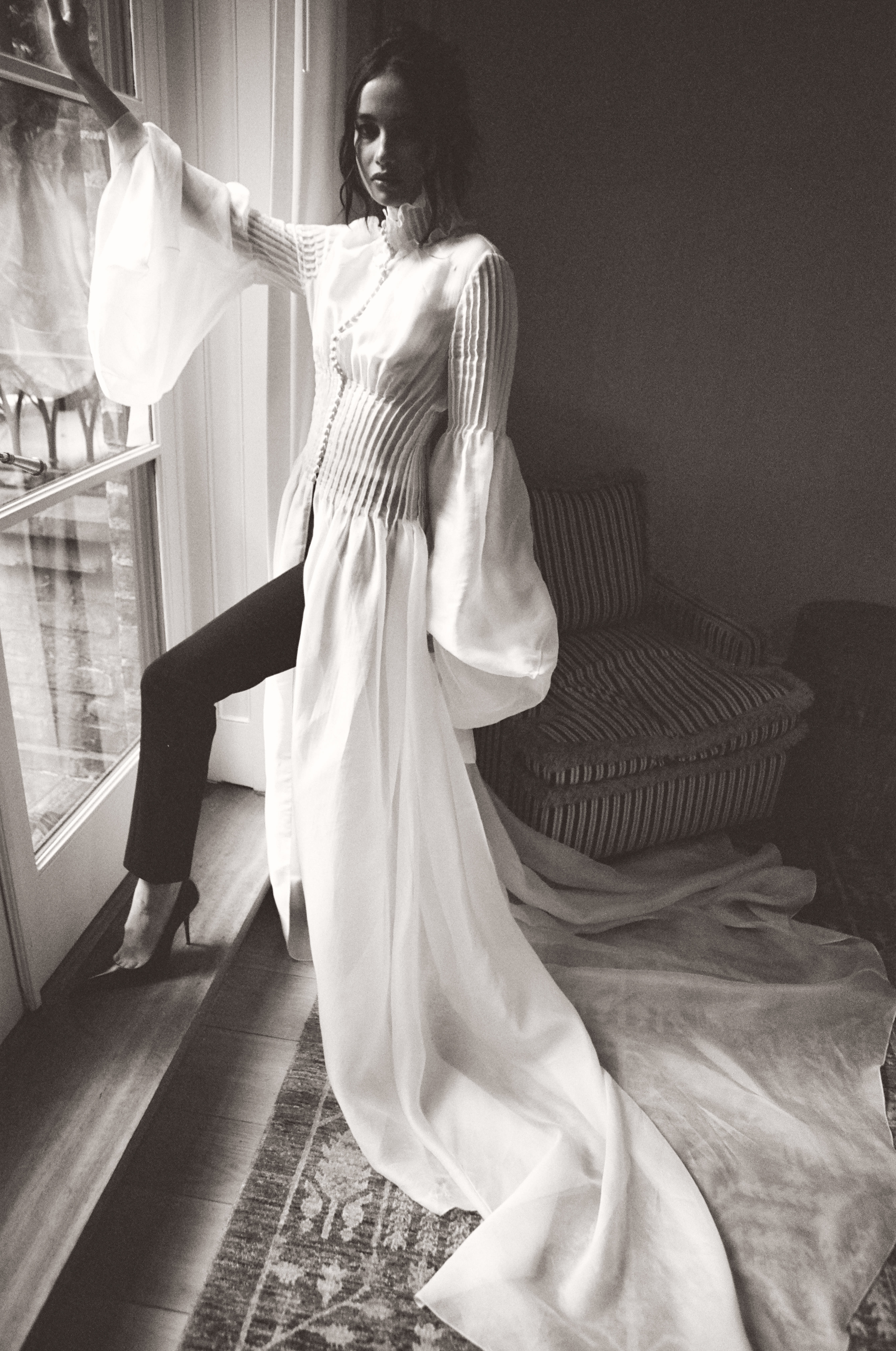 Black and white portrait woman by window in flowing white pleated shirt with pearl necklace