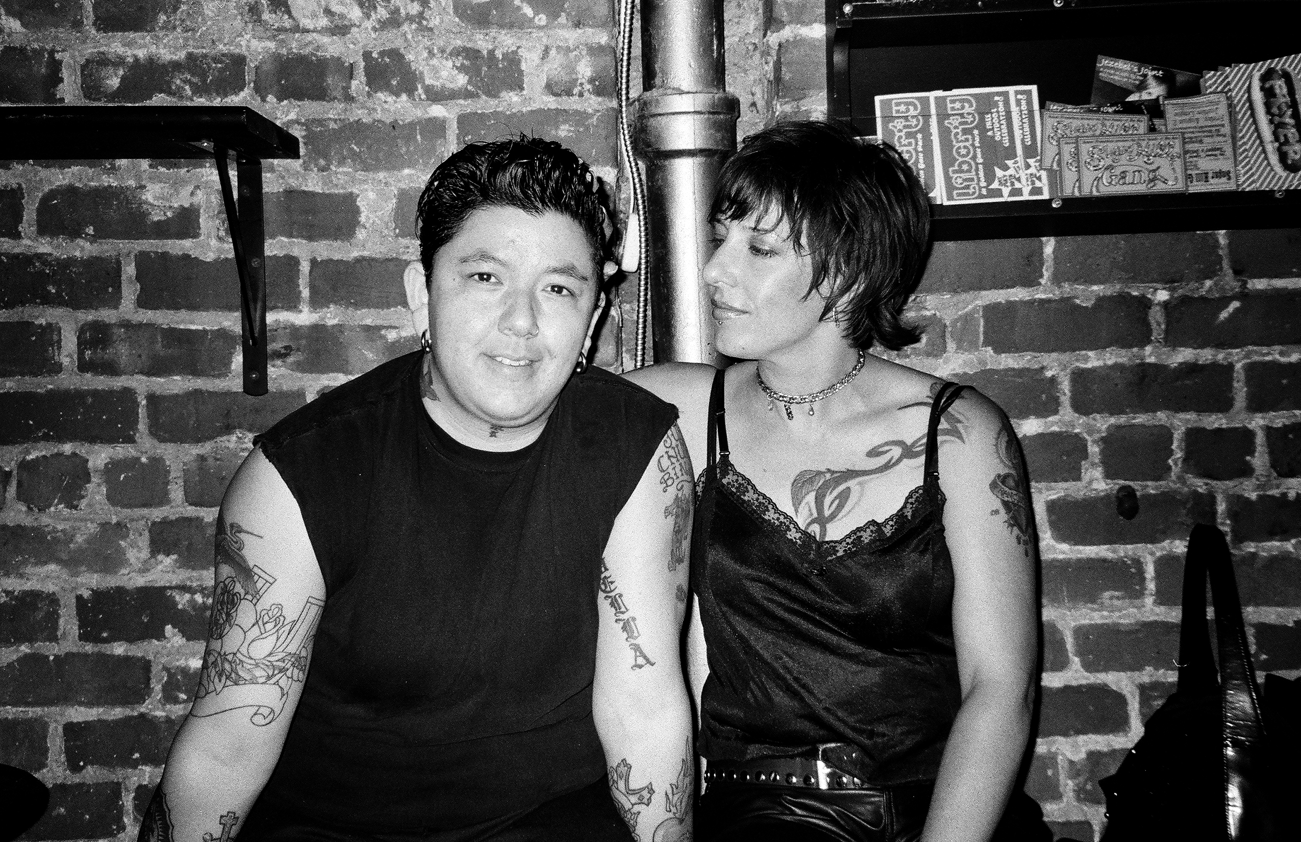 Two tattooed individuals in black clothing against brick wall in intimate bar setting