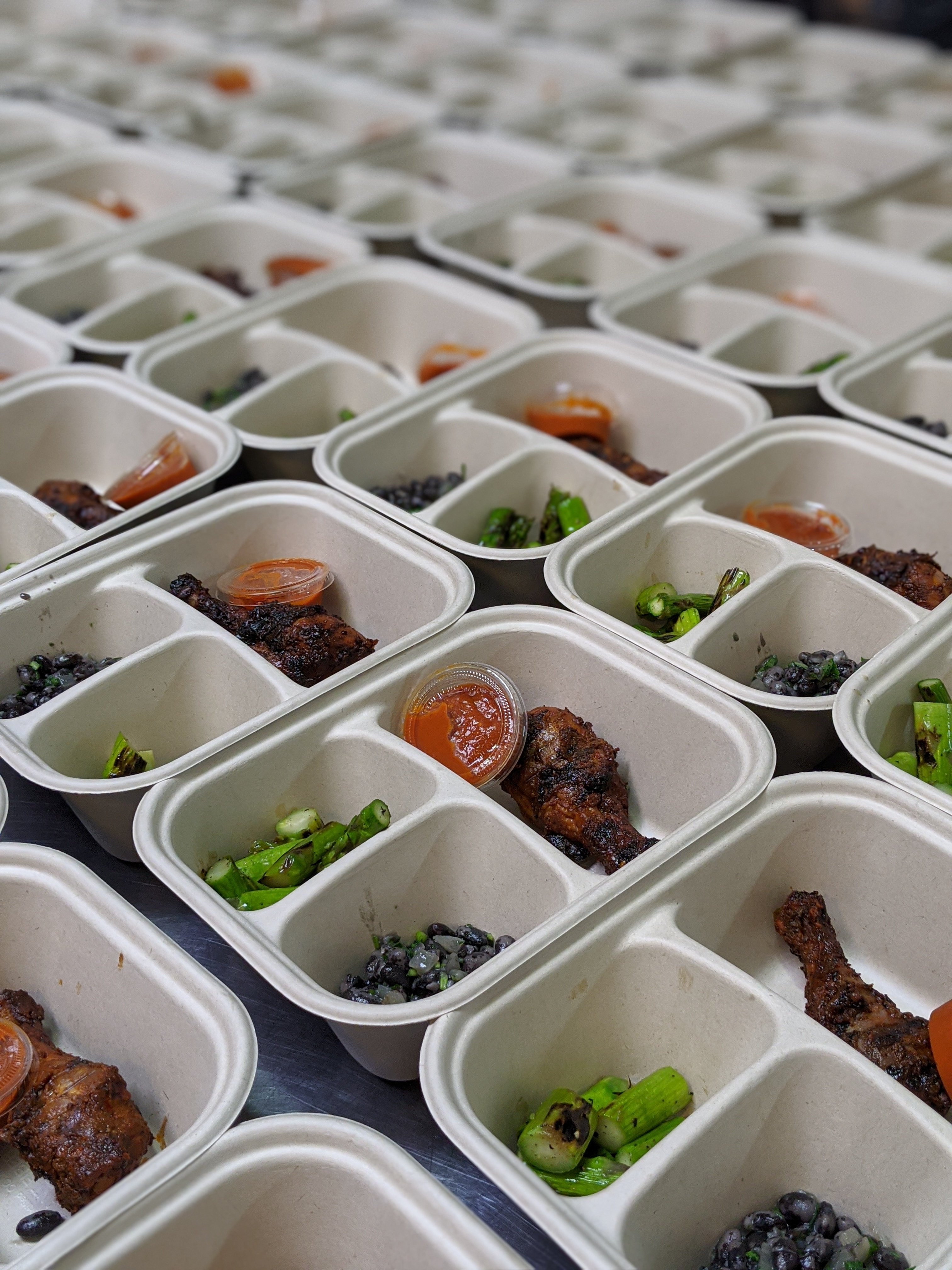 Rows of compostable meal trays filled with jerk chicken, black beans, asparagus, and sauce