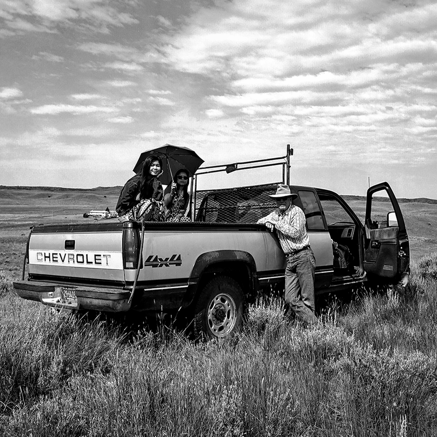 People in Chevrolet 4x4 pickup truck bed with cowboy standing beside parked in open grassland under big sky
