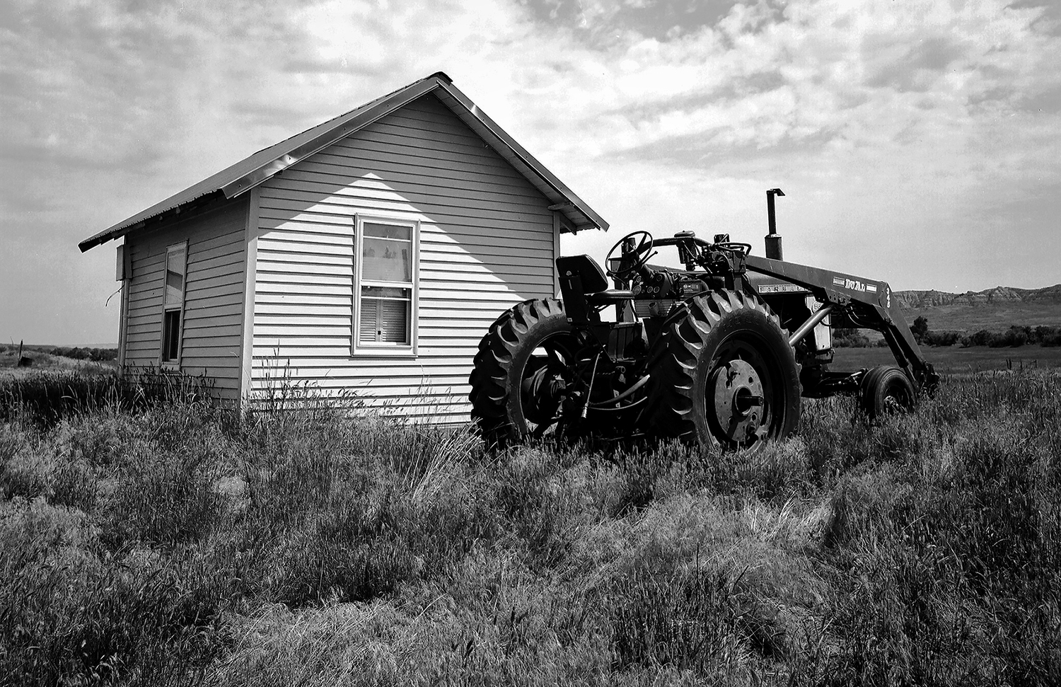 Small white farmhouse with tractor and front loader parked alongside in prairie grassland with badlands in distance