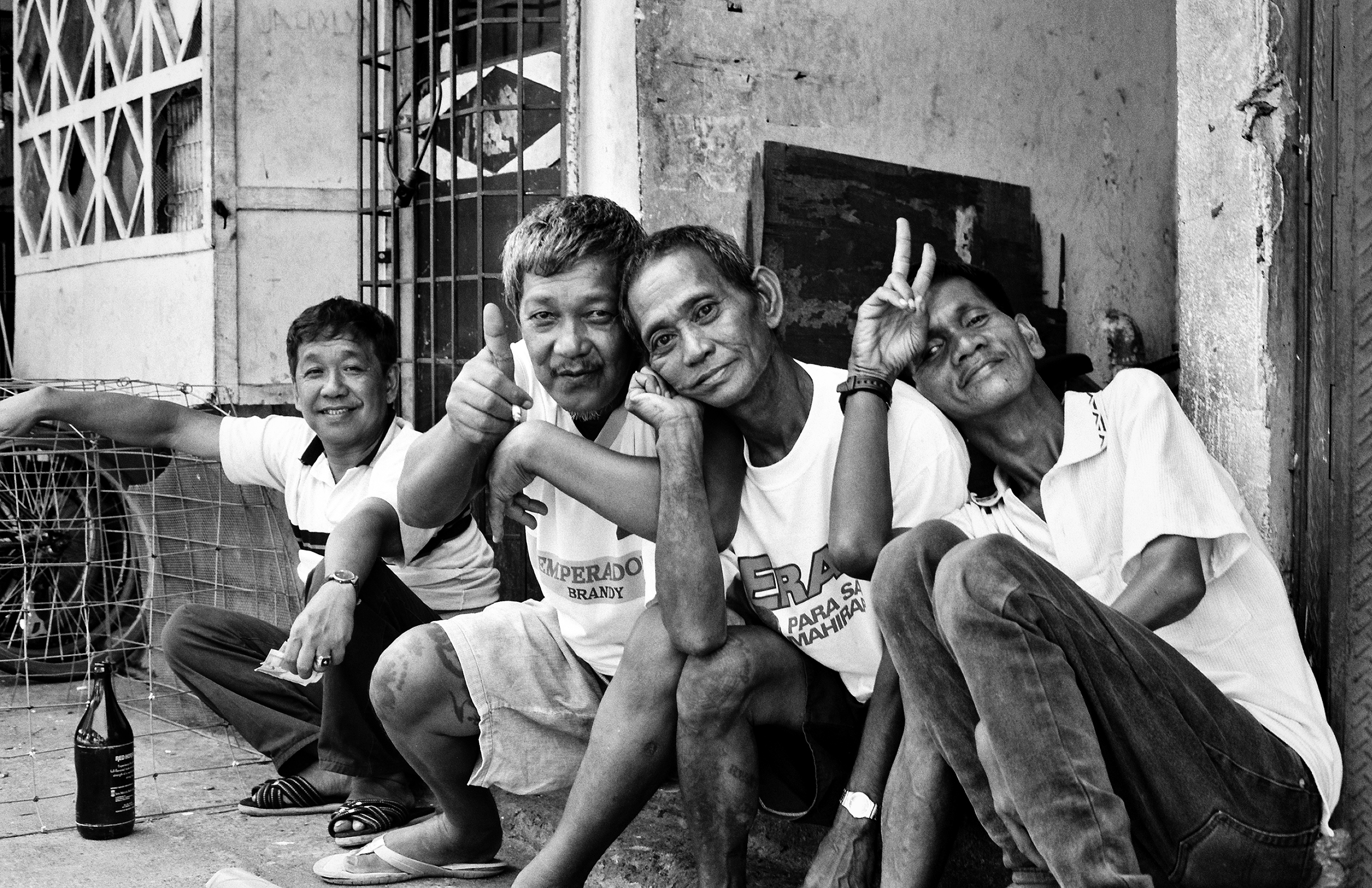 Four middle-aged men sitting together on street smiling and giving thumbs up in candid moment with bottle nearby