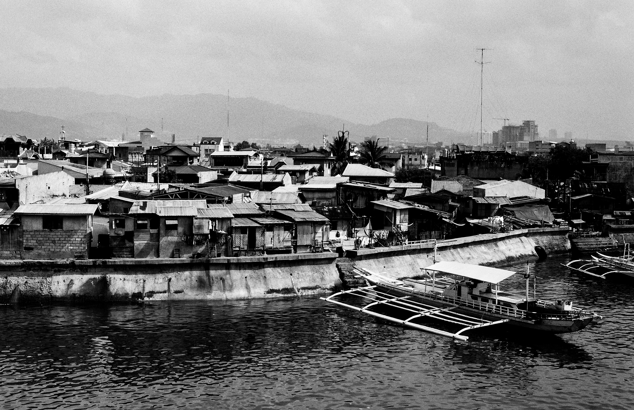 Waterfront view of densely packed residential buildings on seawall with traditional bangka boats moored in foreground and mountains in background